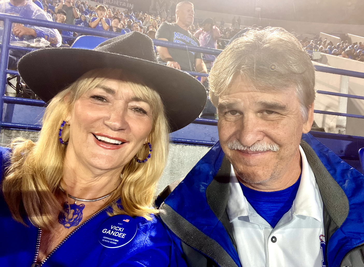 A woman with blonde hair wearing a black hat, blue jacket, and a campaign button that reads 'Vicki Gandee, Germantown School Board.' She is smiling. A man with gray hair and a mustache, wearing a blue and white jacket, is also smiling. They are at a crowded outdoor event, possibly a sports game, with blue seats and other attendees in the background.