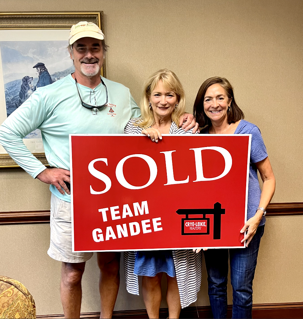 Three people, two women and one man, smiling and holding a red sign that says "SOLD TEAM GANDEE" with a logo for Crye-Leike Realtors. They are indoors against a beige wall with a framed picture of dogs in the background.
