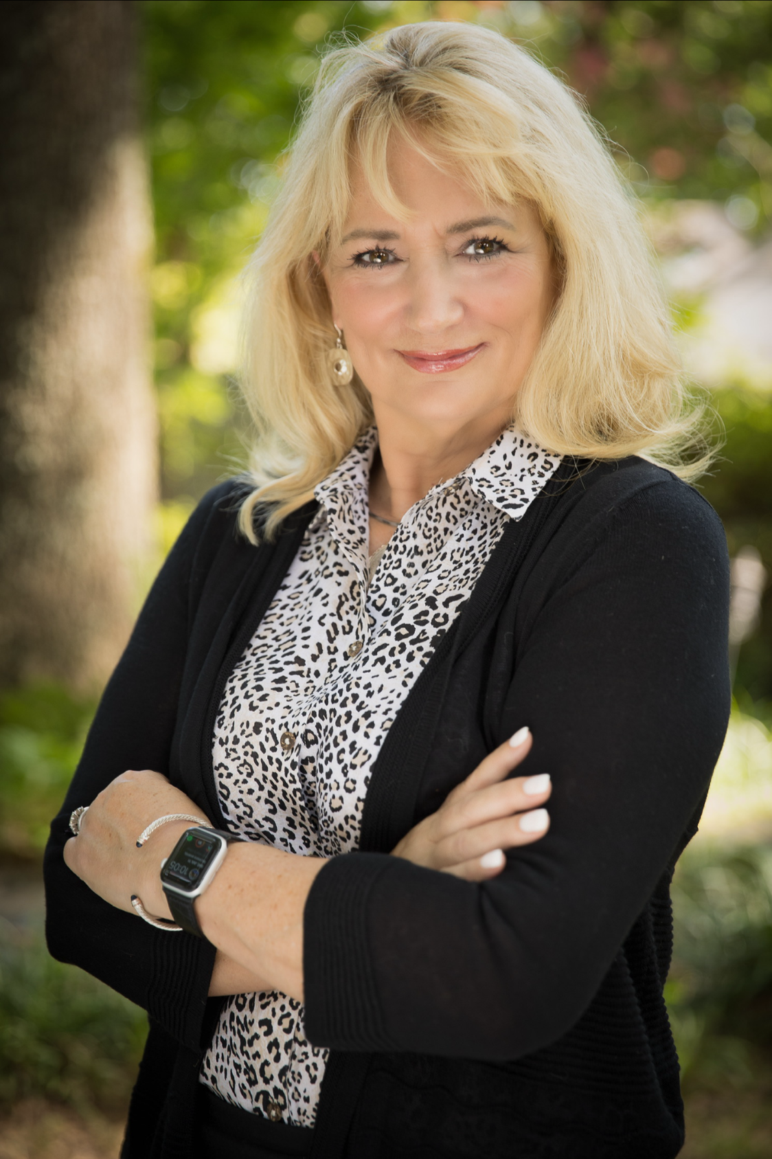 A woman with blonde hair and light skin standing outdoors with her arms crossed, smiling, wearing a black cardigan over a leopard print blouse and a smartwatch on her wrist, with green trees and sunlight in the background.