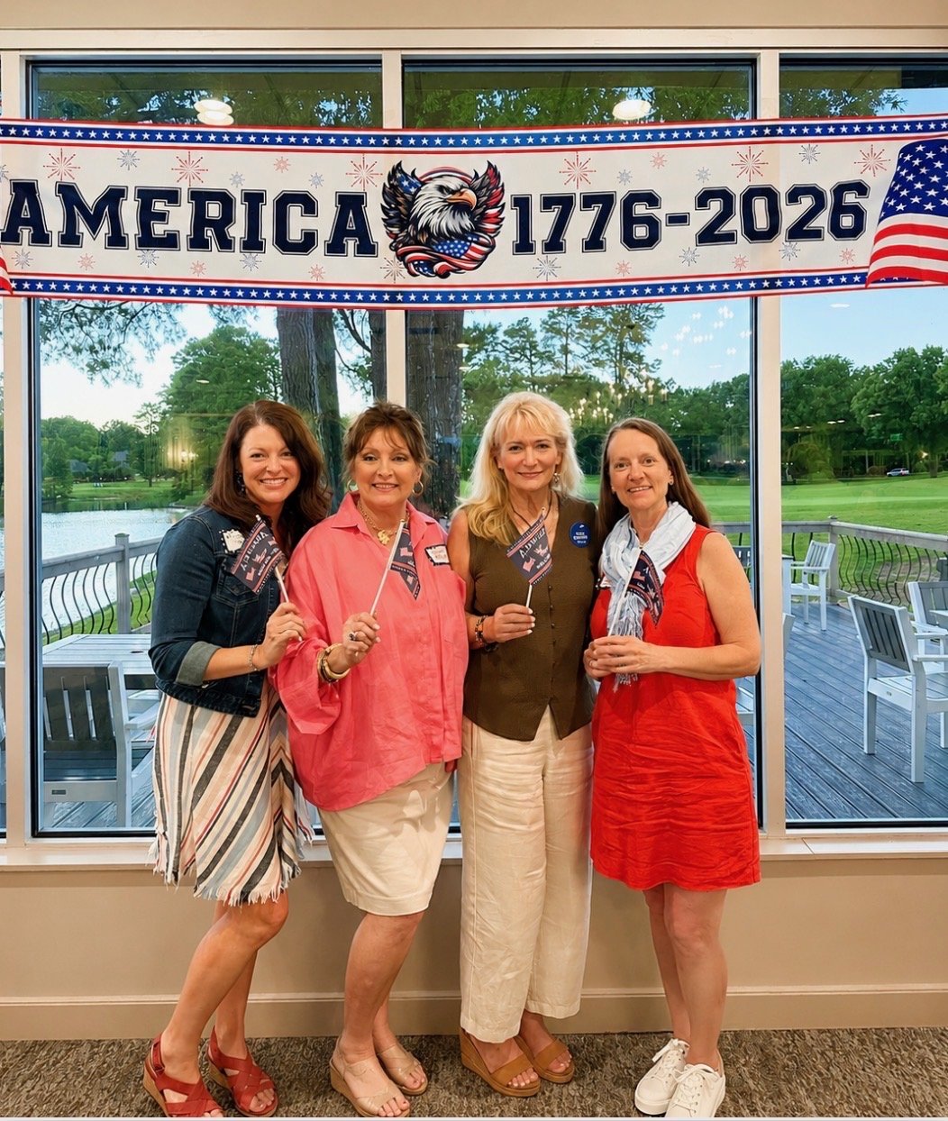 Four women standing in front of a large window with a scenic view of a park and water, holding small American flags, under a patriotic banner that reads 'America 1776-2026' with an eagle illustration, celebrating a patriotic event or anniversary.