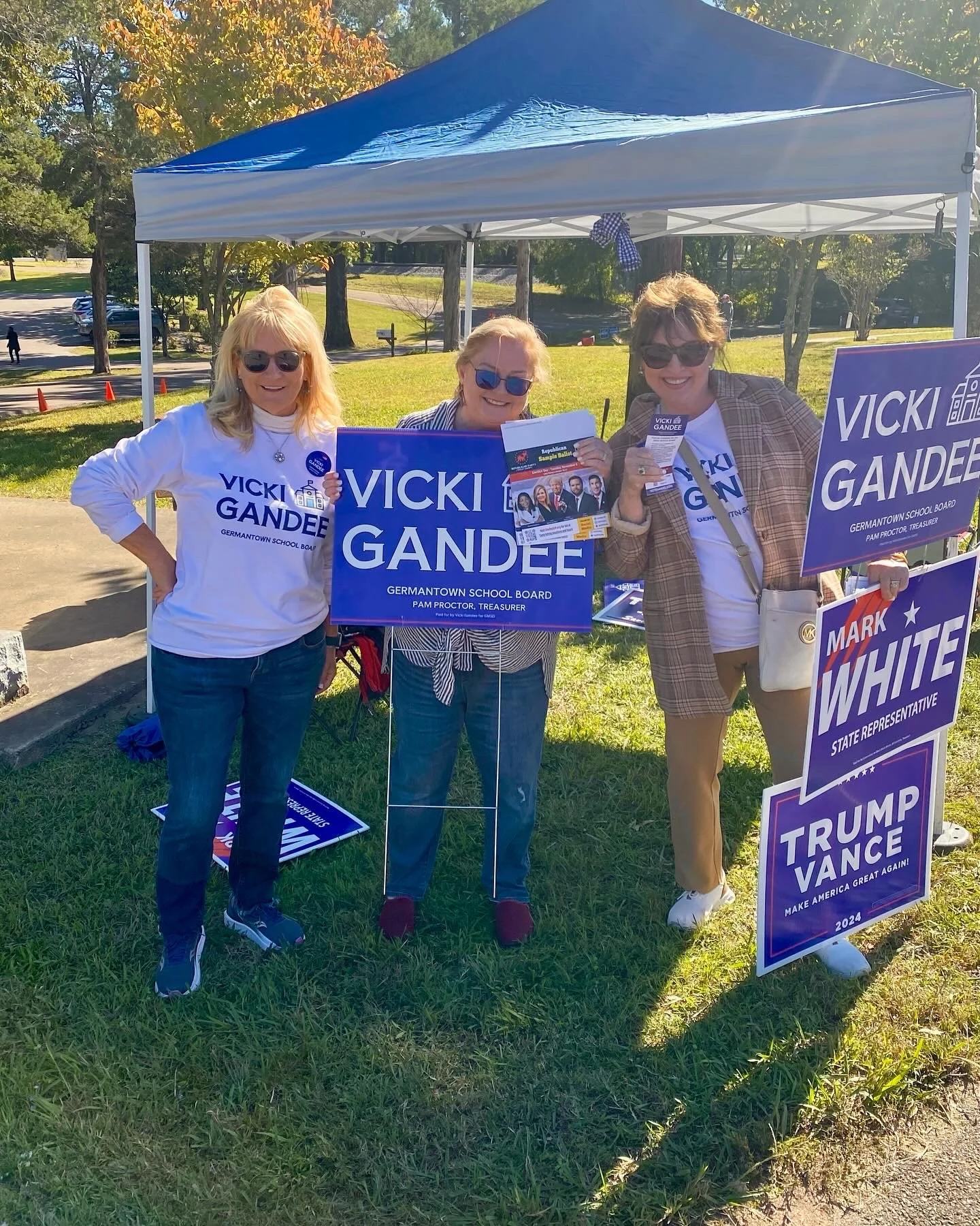 Three women standing under a blue canopy at a political event outdoors, holding and displaying campaign signs for Vicki Gandee and Trump Vance, with trees and a grassy area in the background.