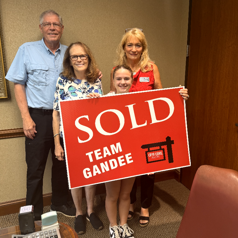 Four people standing indoors, holding a large red 'SOLD' sign with 'TEAM GANDEE' and a logo, smiling at the camera. One person is a young girl with a ponytail, and the others are adults, including an older man and woman, and another woman with glasses and blonde hair.