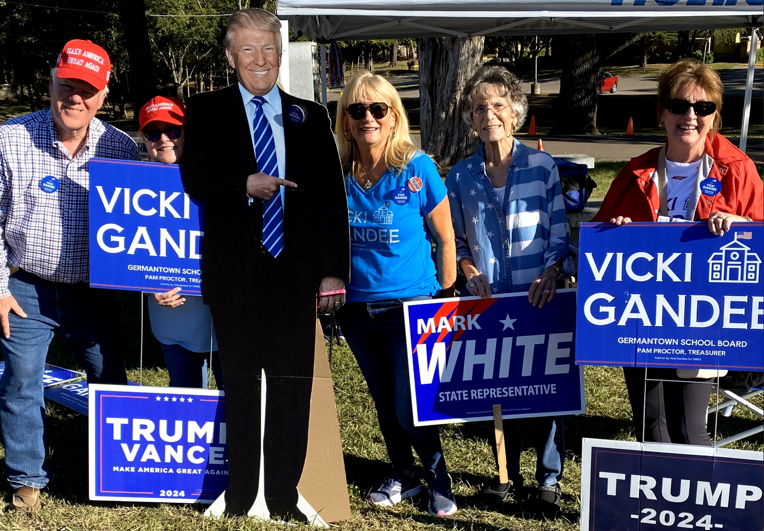 Group of people standing outdoors holding political campaign signs, including one of Donald Trump and several supporting Vicki Gander and Mark White in 2024, with one person dressed as a cardboard cutout of Donald Trump.