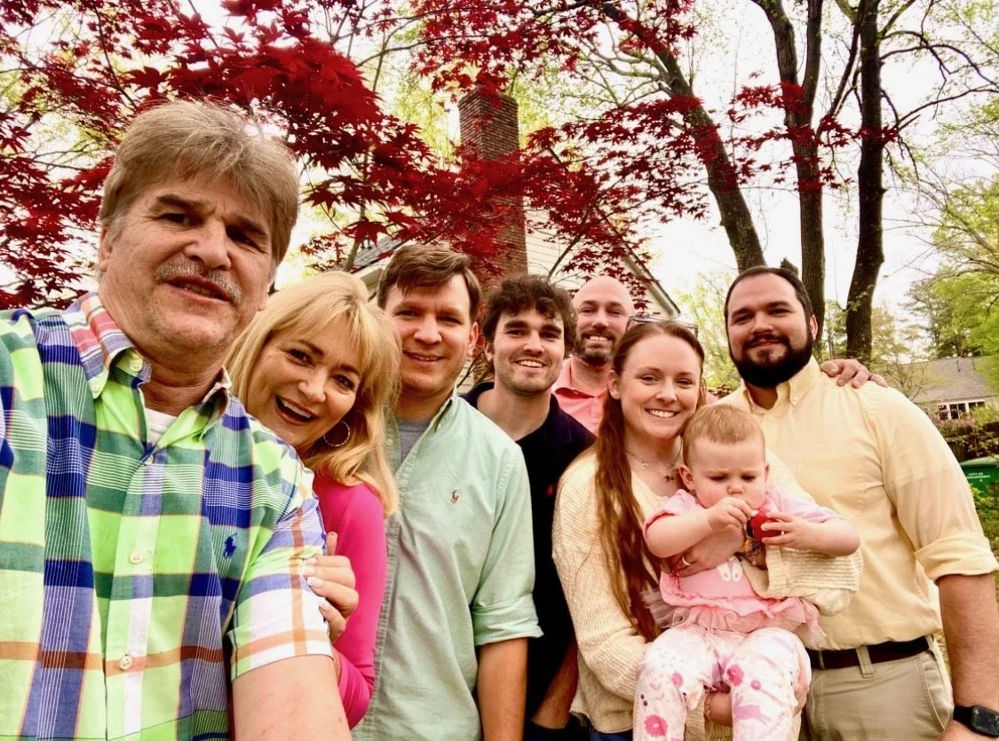 Group of nine people, including children, smiling outdoors on a sunny day with trees and a house in the background.