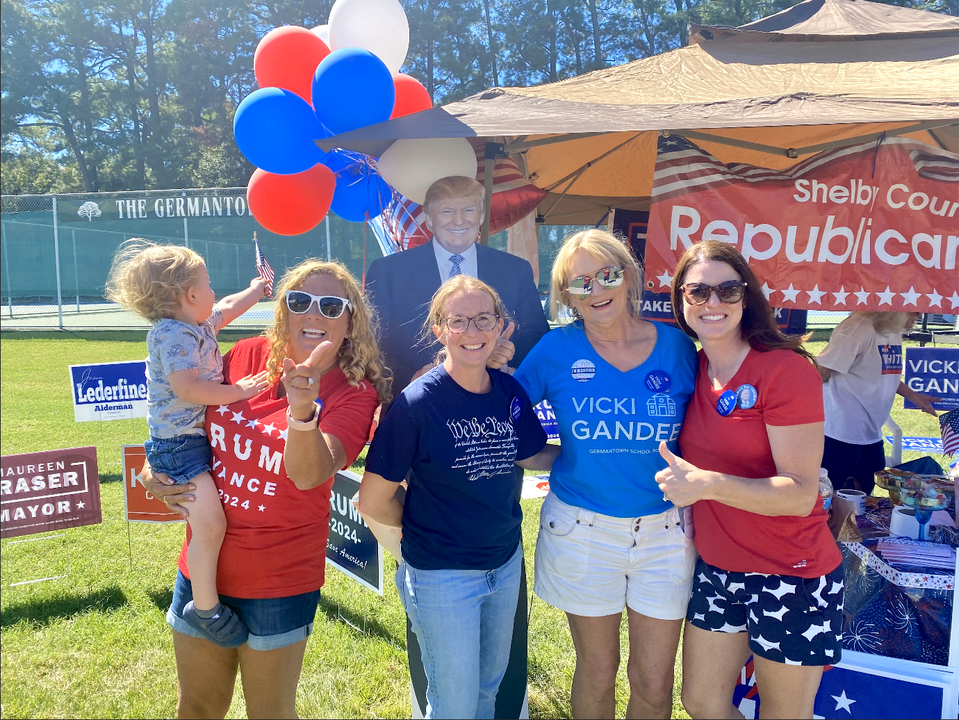 Group of women and a child at a political rally with red, white, and blue balloons, campaign signs for Donald Trump and Vicki Gande, and a cardboard cutout of Donald Trump. They are outdoors on a grassy field with tennis courts and trees in the background.
