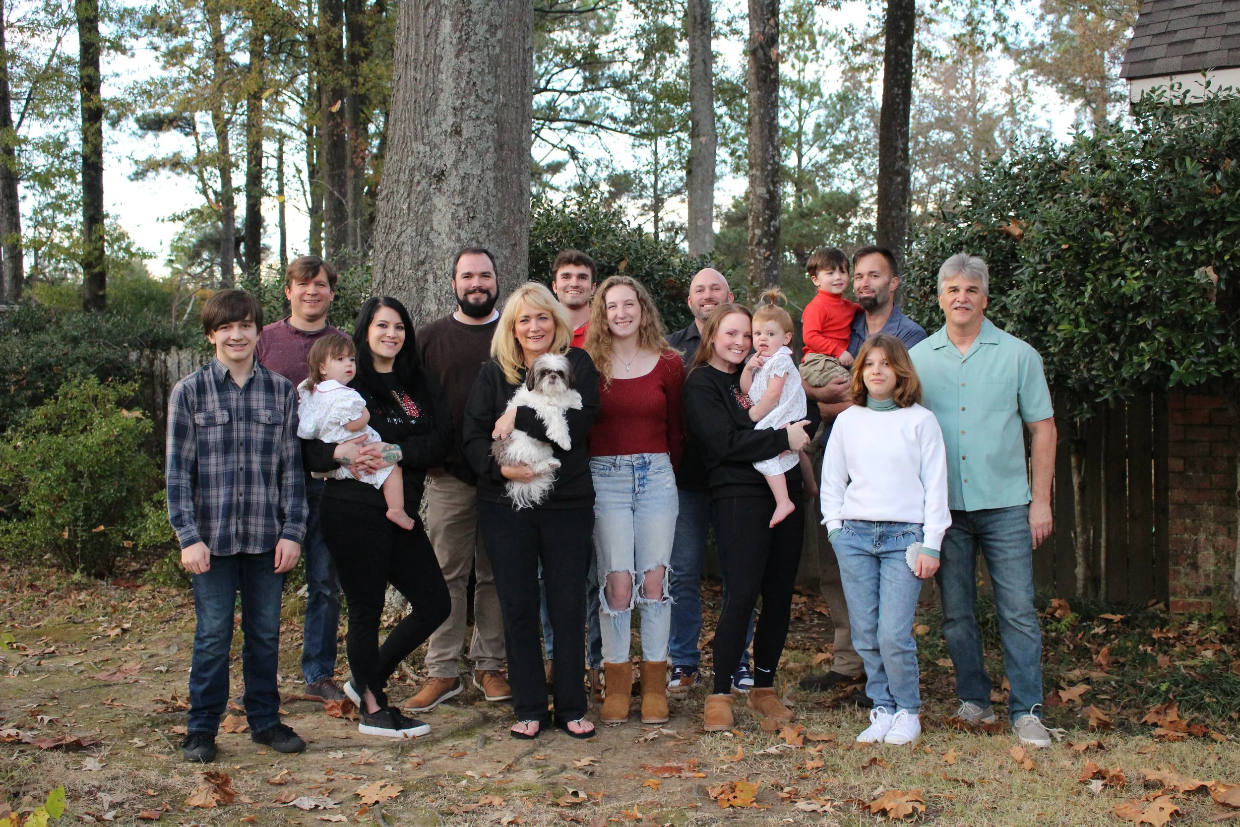 A multi-generational family group photo outdoors in a wooded area, including children, teenagers, adults, and an elderly man, some holding young children and a small dog, all smiling at the camera.