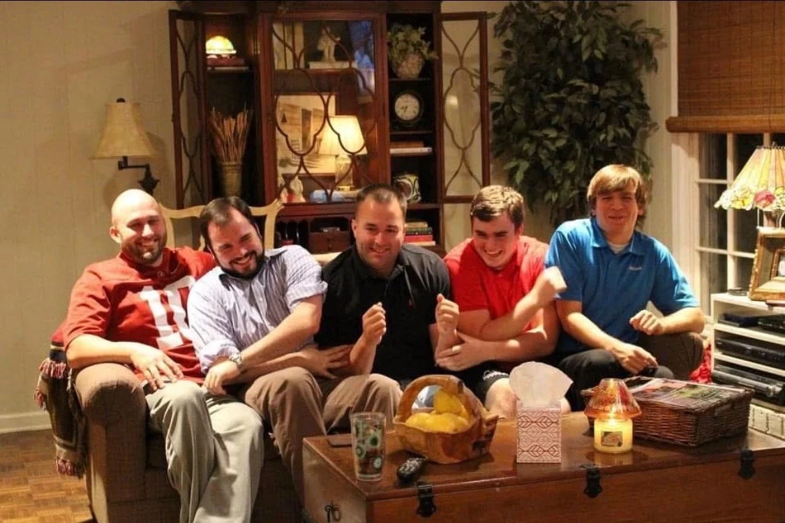 Five young men sitting on a beige sofa in a living room, smiling and posing for the photo. They are dressed casually, and there are various decorative items on a wooden cabinet and a coffee table in front of them.