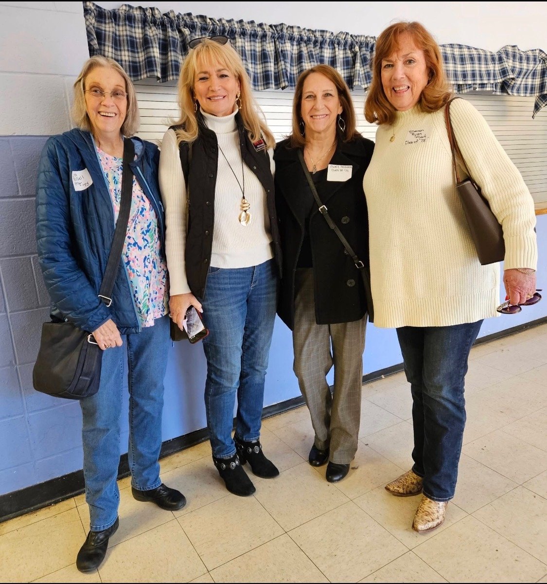 Four women standing together indoors, smiling, wearing casual to business casual clothing, in front of a window with a checkered valance, at an event or gathering.