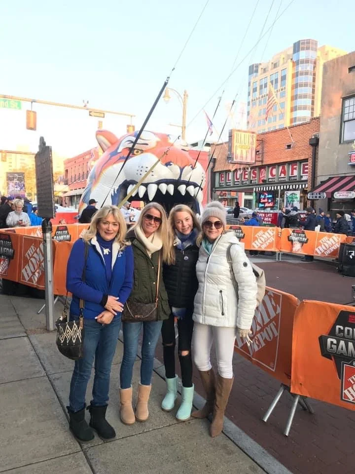 Four women standing on a sidewalk in front of an amusement ride and a blues café, with other people and barricades behind them.
