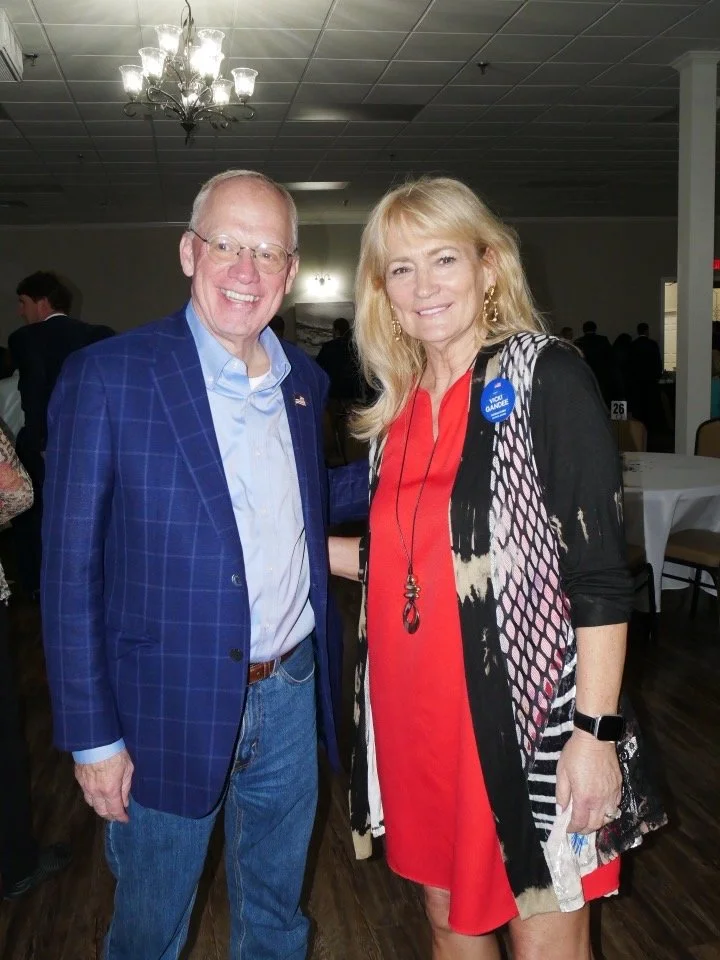 A man in a blue checked blazer and a woman in a red dress and black-and-white patterned cardigan standing together at an indoor event, smiling at the camera.