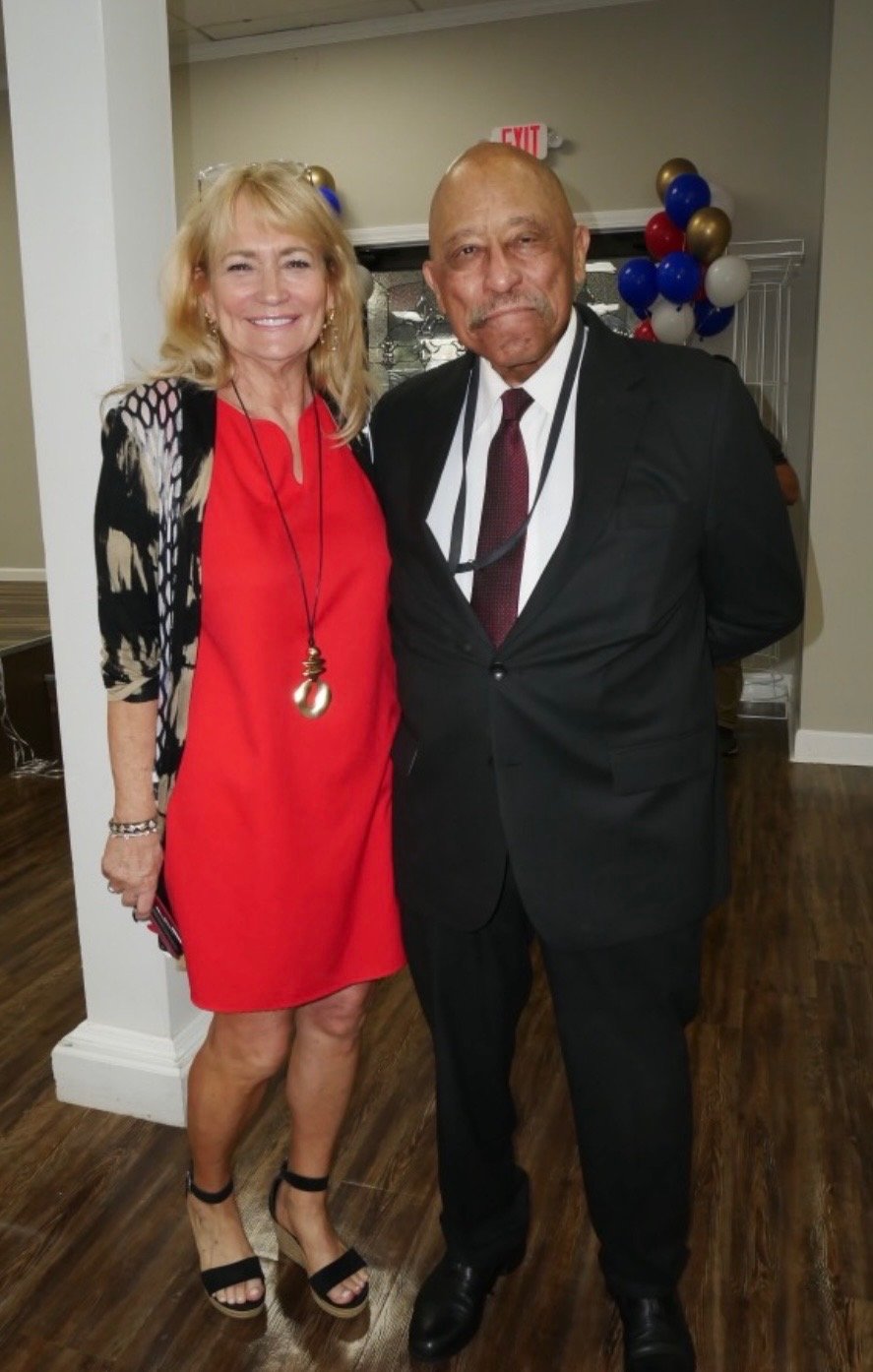 A woman and a man stand together inside a decorated room, smiling at the camera. The woman is wearing a red dress with a black and white patterned cardigan and black high heels. The man is dressed in a black suit, white shirt, and maroon tie.