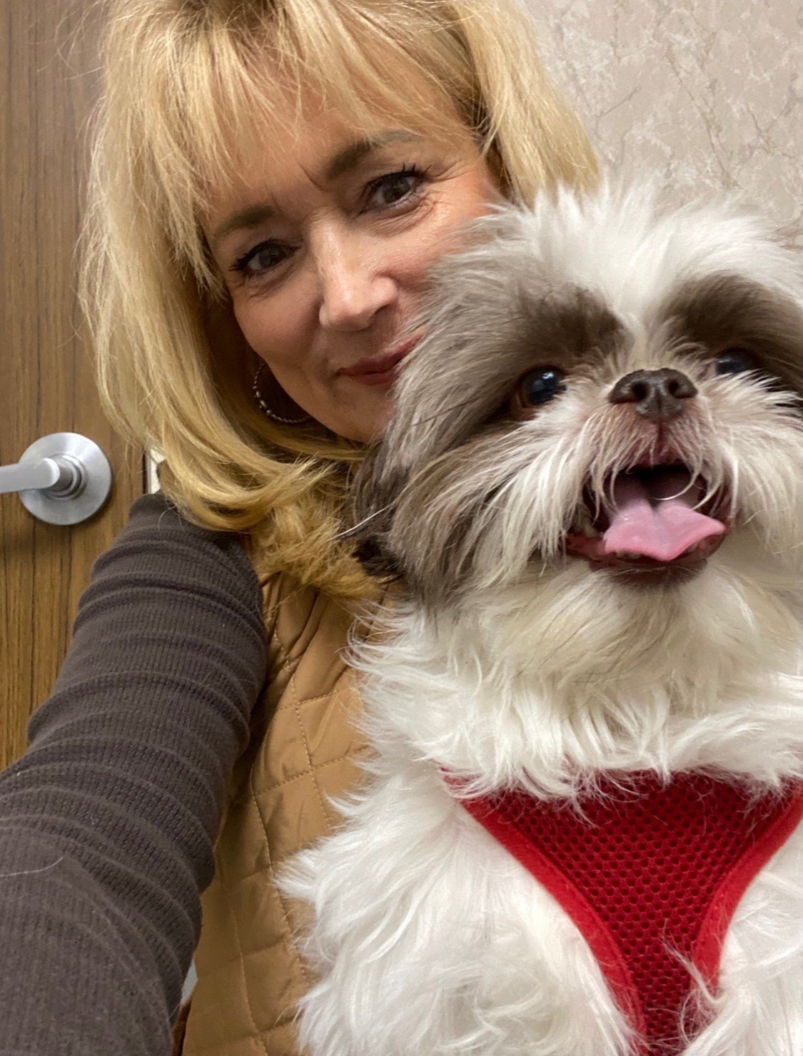 A woman with blonde hair smiling next to a fluffy, happy dog with a red harness.