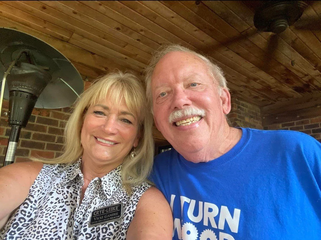 A smiling woman and man taking a selfie together inside a wooden-paneled room, with a brick wall and heater in the background. The woman is wearing a sleeveless blouse with a leopard pattern and a name tag, and the man is wearing a bright blue T-shir