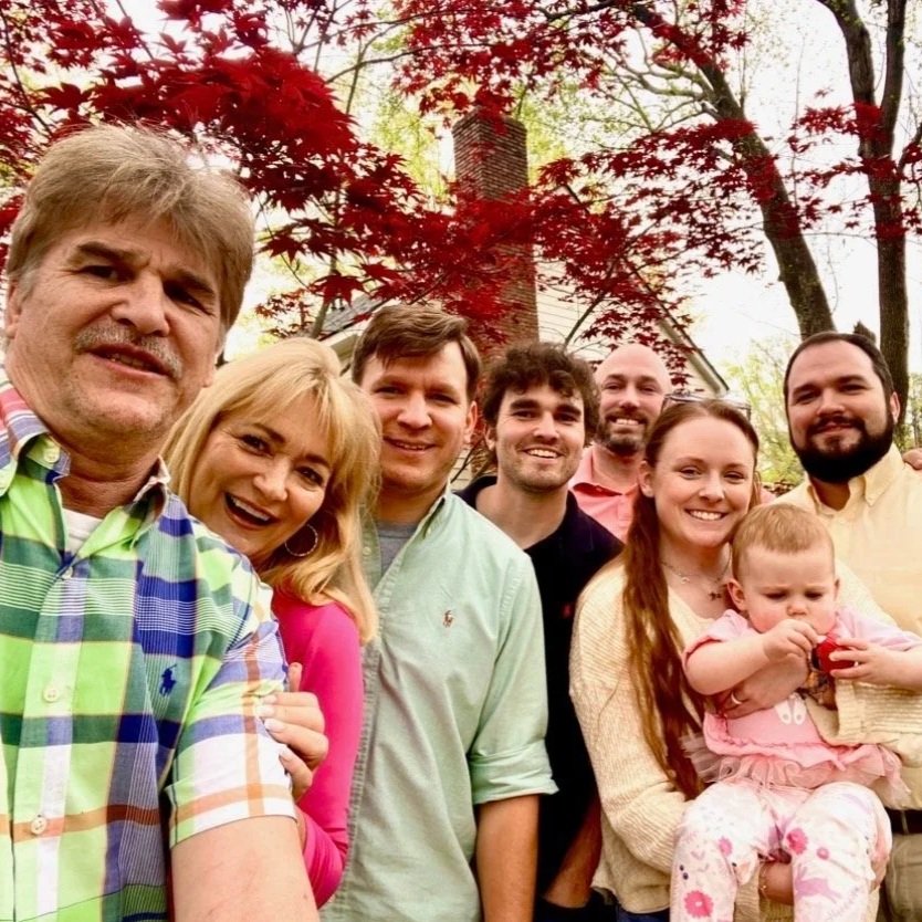 Group of seven people, including a child, standing outdoors in front of trees with red leaves, smiling at the camera.