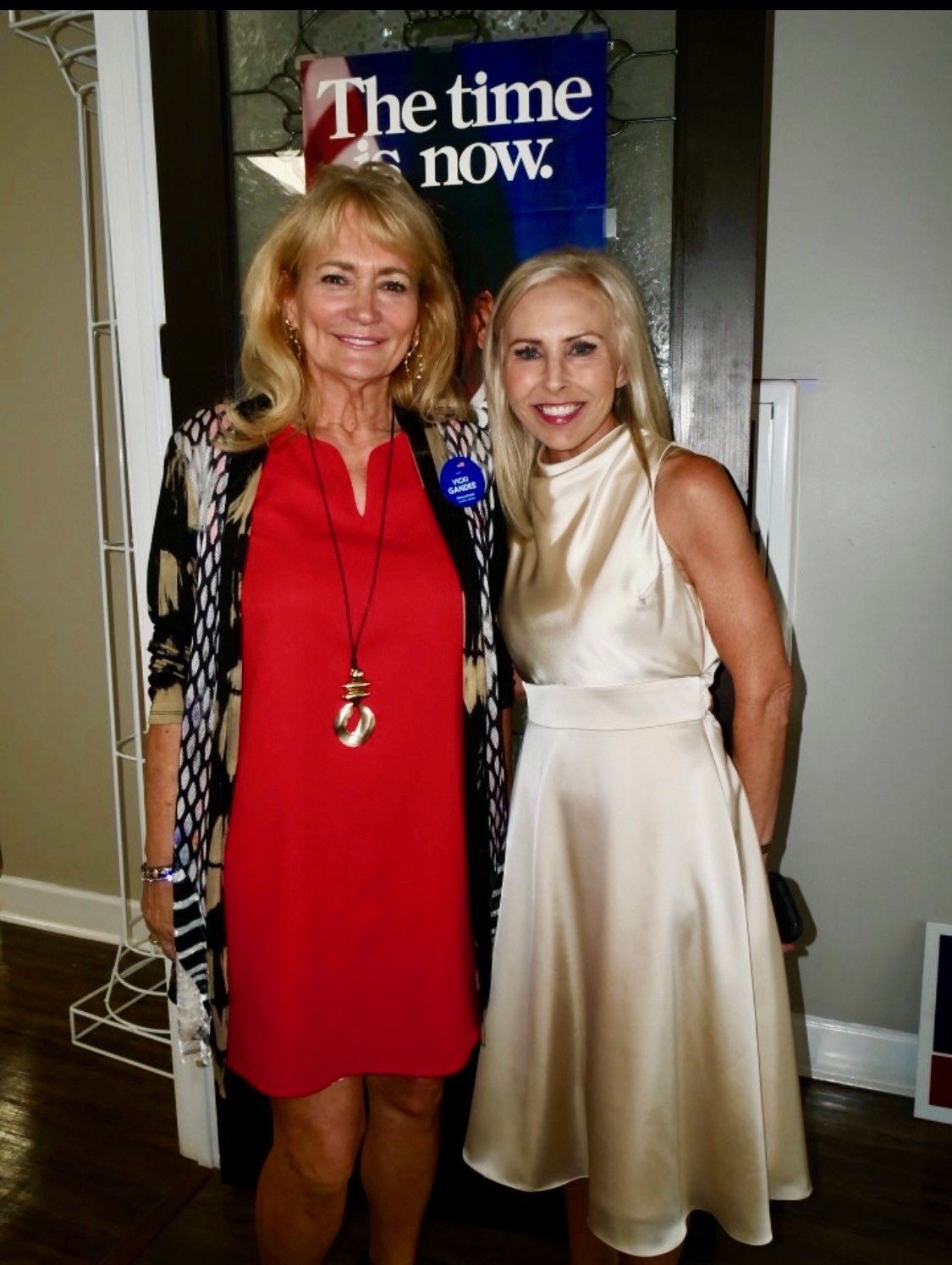 Two women smiling and standing close together inside, one wearing a red dress with a black and white patterned cardigan, and the other in a cream-colored satin dress, with a sign in the background that says "The time is now."