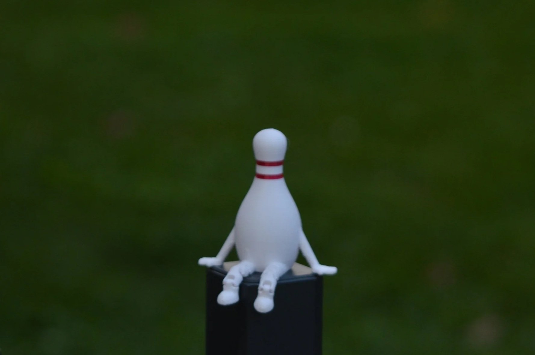 A ceramic figurine of a person with a bowling pin for a head, sitting on a black box with arms and legs extended, against a blurred green background.
