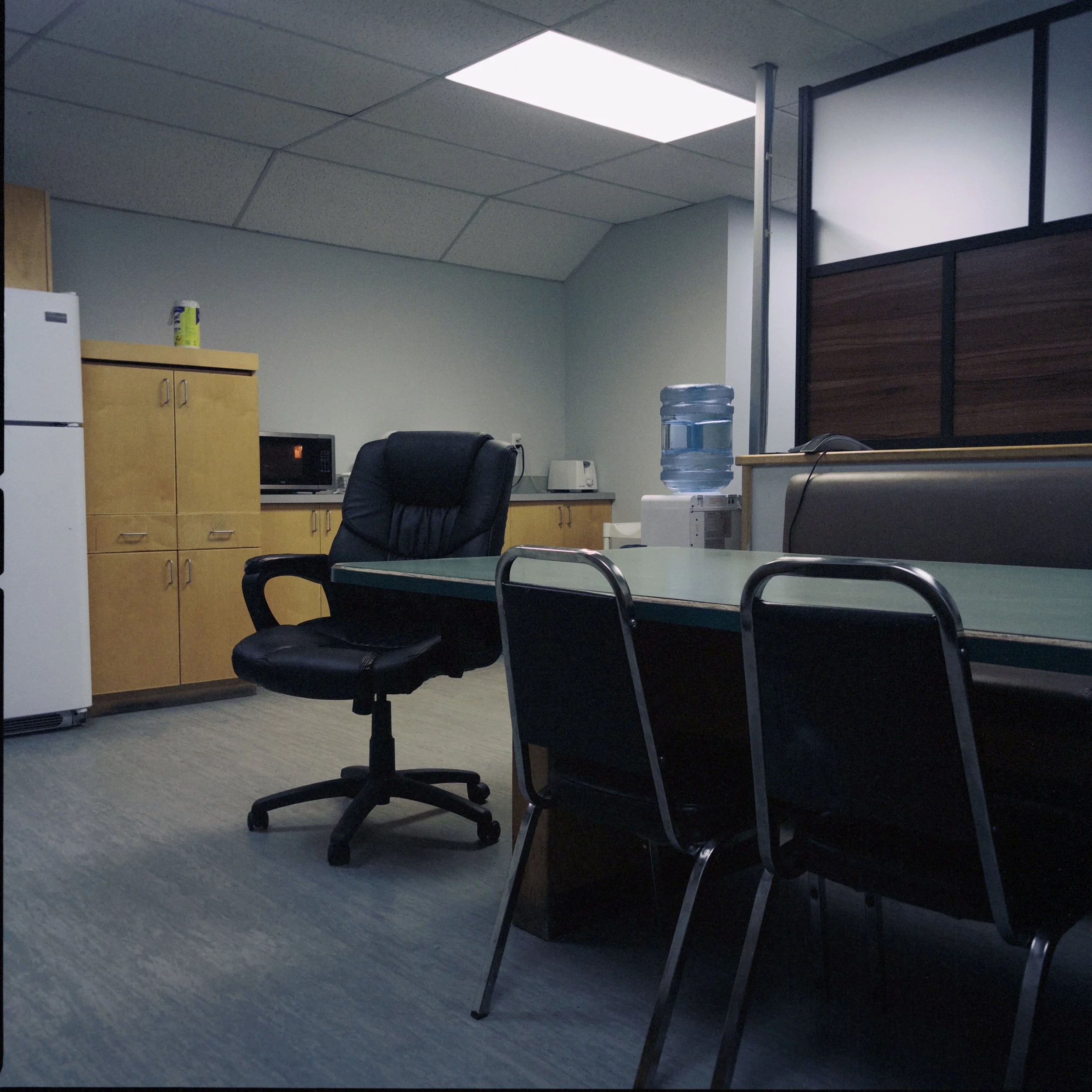 An office breakroom with a water cooler, a black office chair, a microwave, a refrigerator, cabinets, and a large panel in the background.