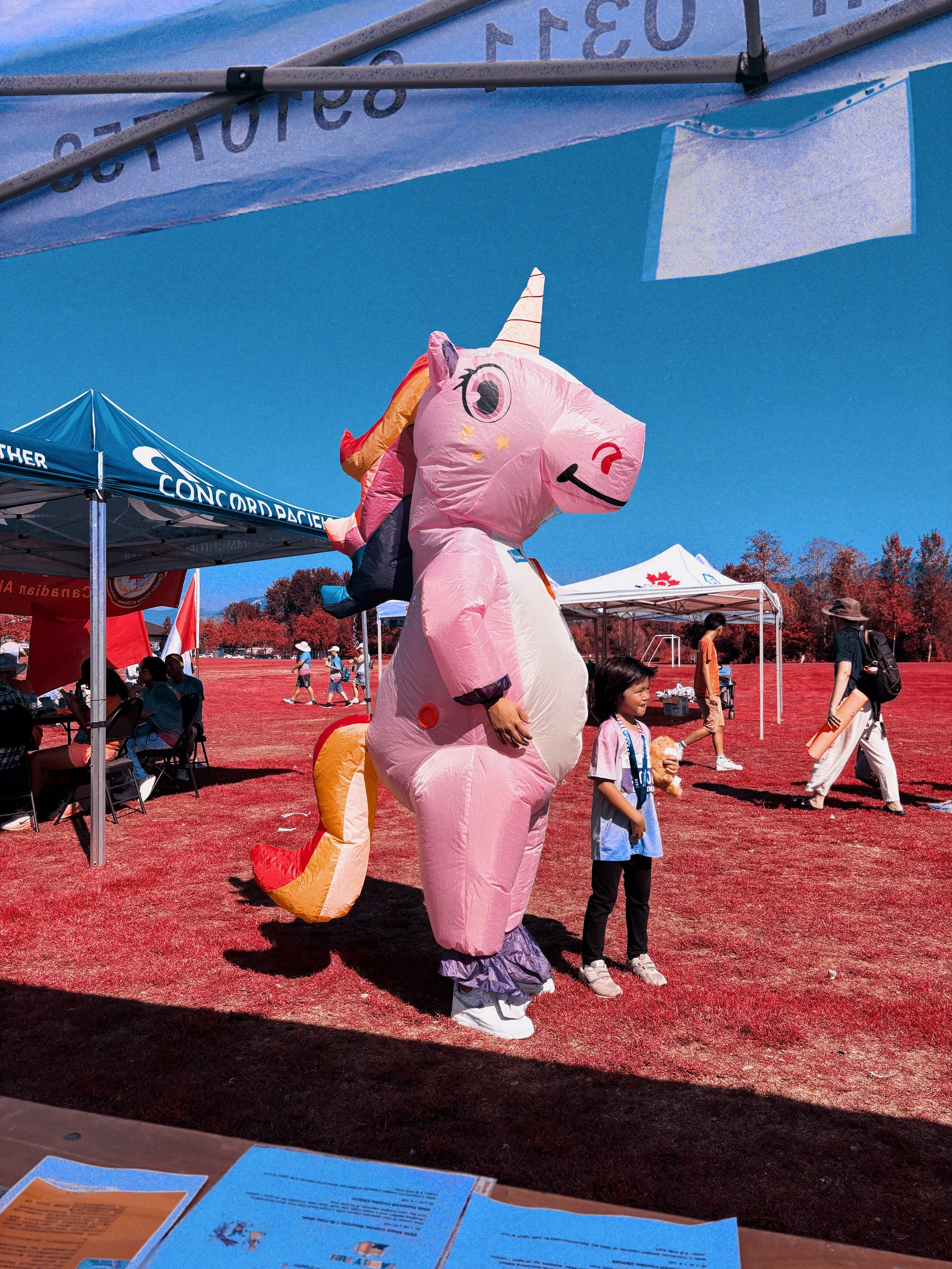 A person dressed in a pink unicorn costume standing on a red sports field surrounded by tents and people at an outdoor event.