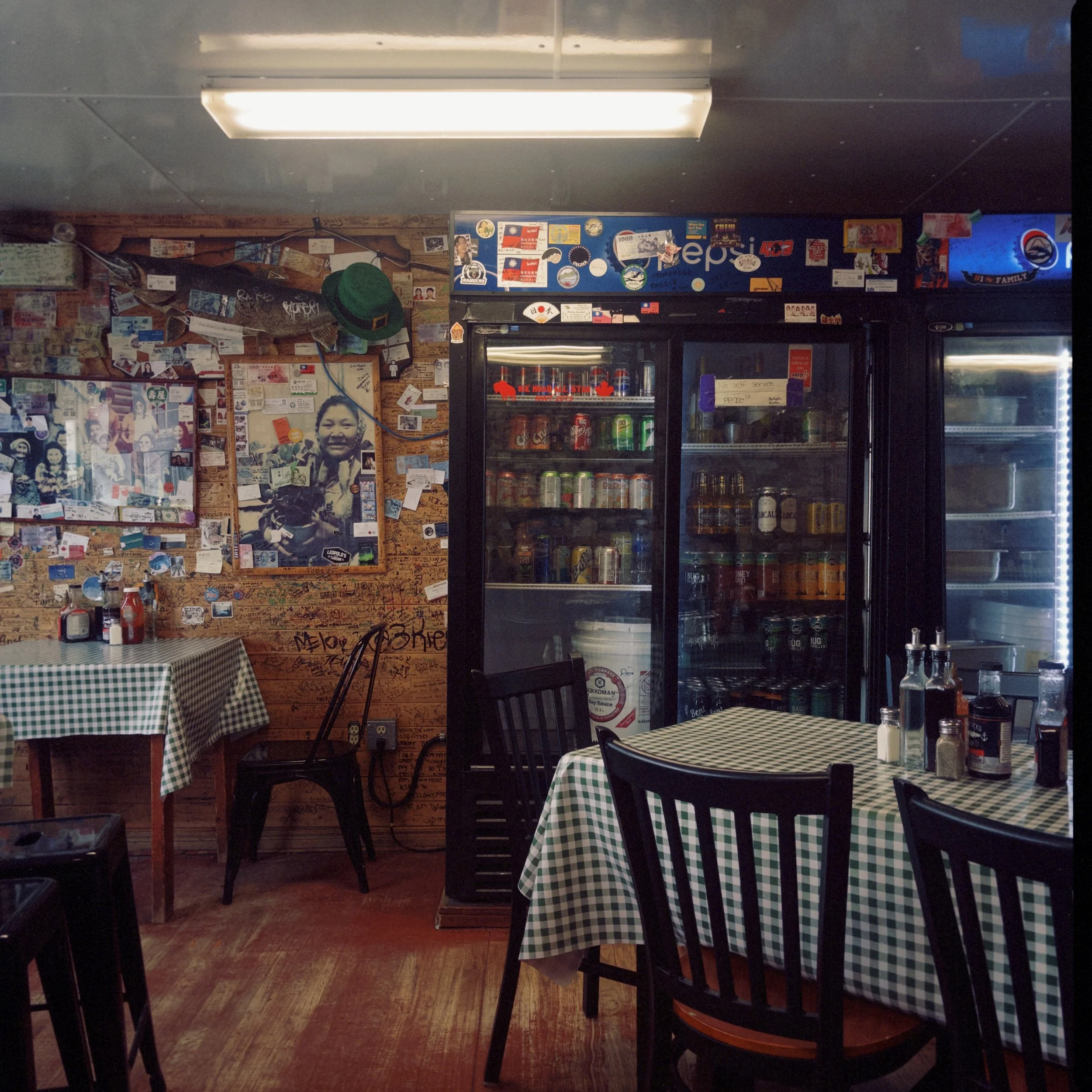 Interior of a small restaurant or cafe with checkered tablecloths on two tables, a brick wall covered in stickers, posters, and graffiti, and a refrigerated display case with soda cans and bottles. A green hat hangs on a hook on the wall.