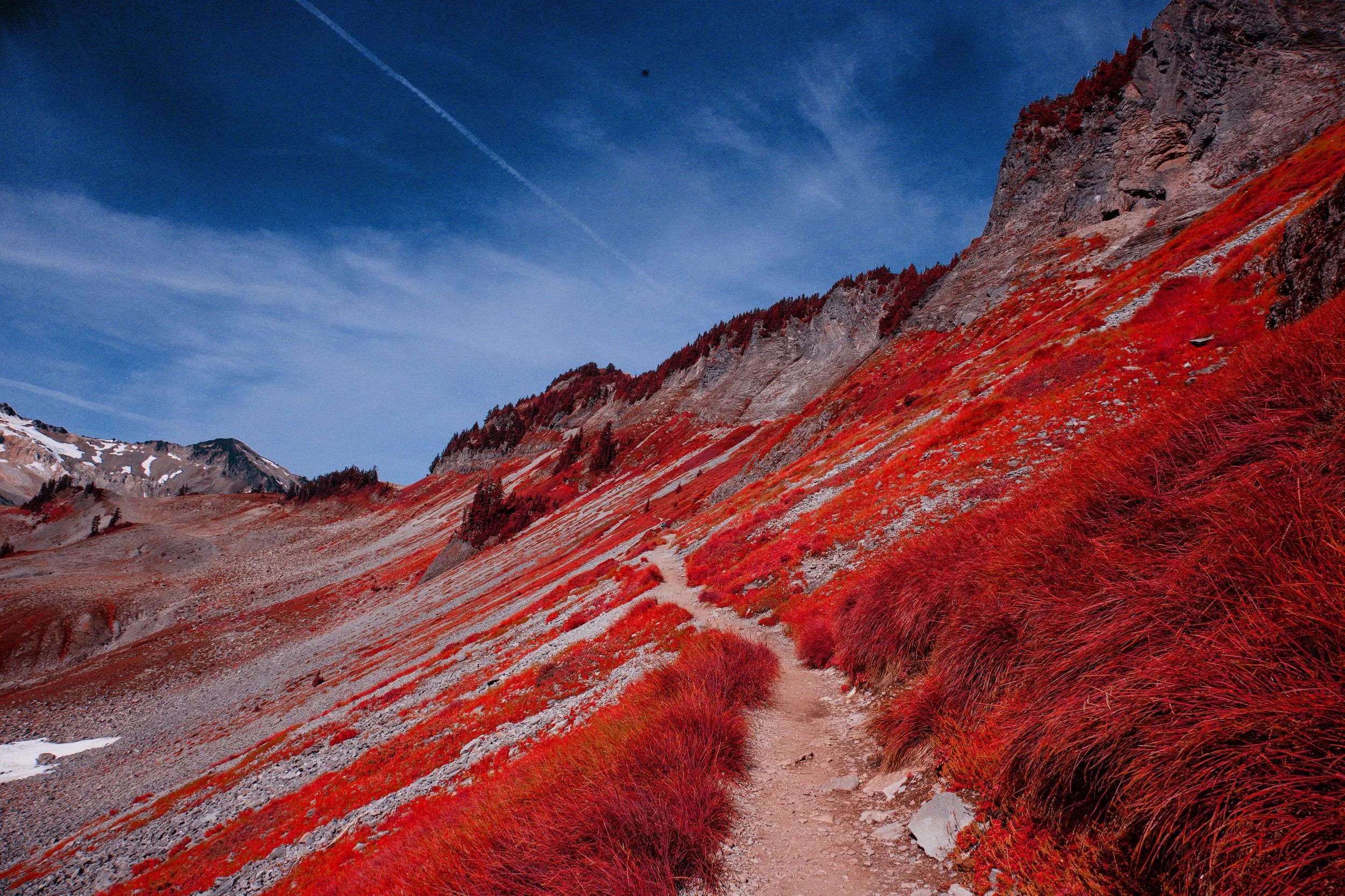 Mountain trail winding through red and green vegetation with rocky slopes and a blue sky with clouds and contrails.