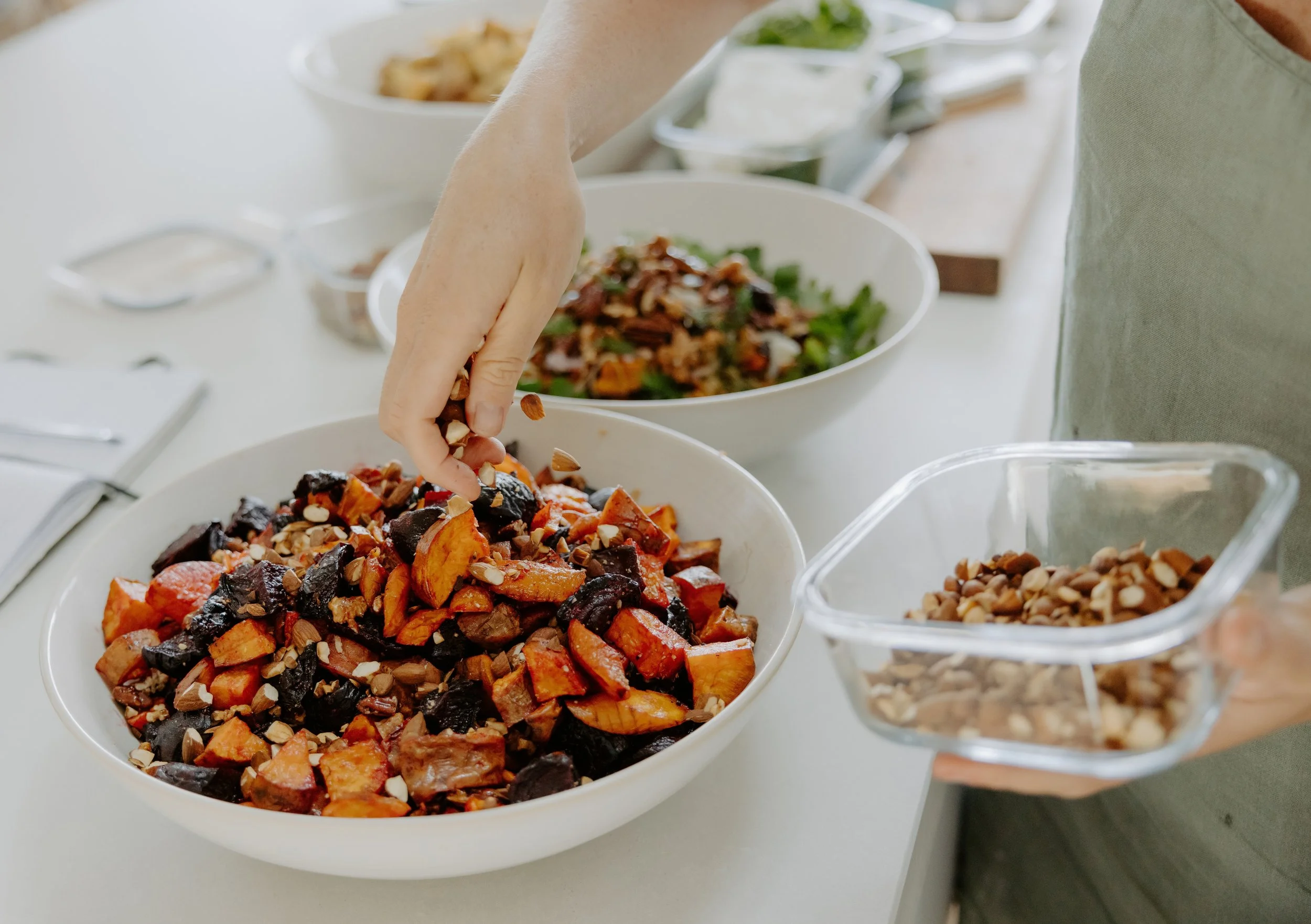 Close-up of a table with various bowls of food, including a bowl of roasted vegetables topped with chopped nuts in the foreground, and a person's hand reaching to pick one. Other bowls in the background contain salads and toppings.
