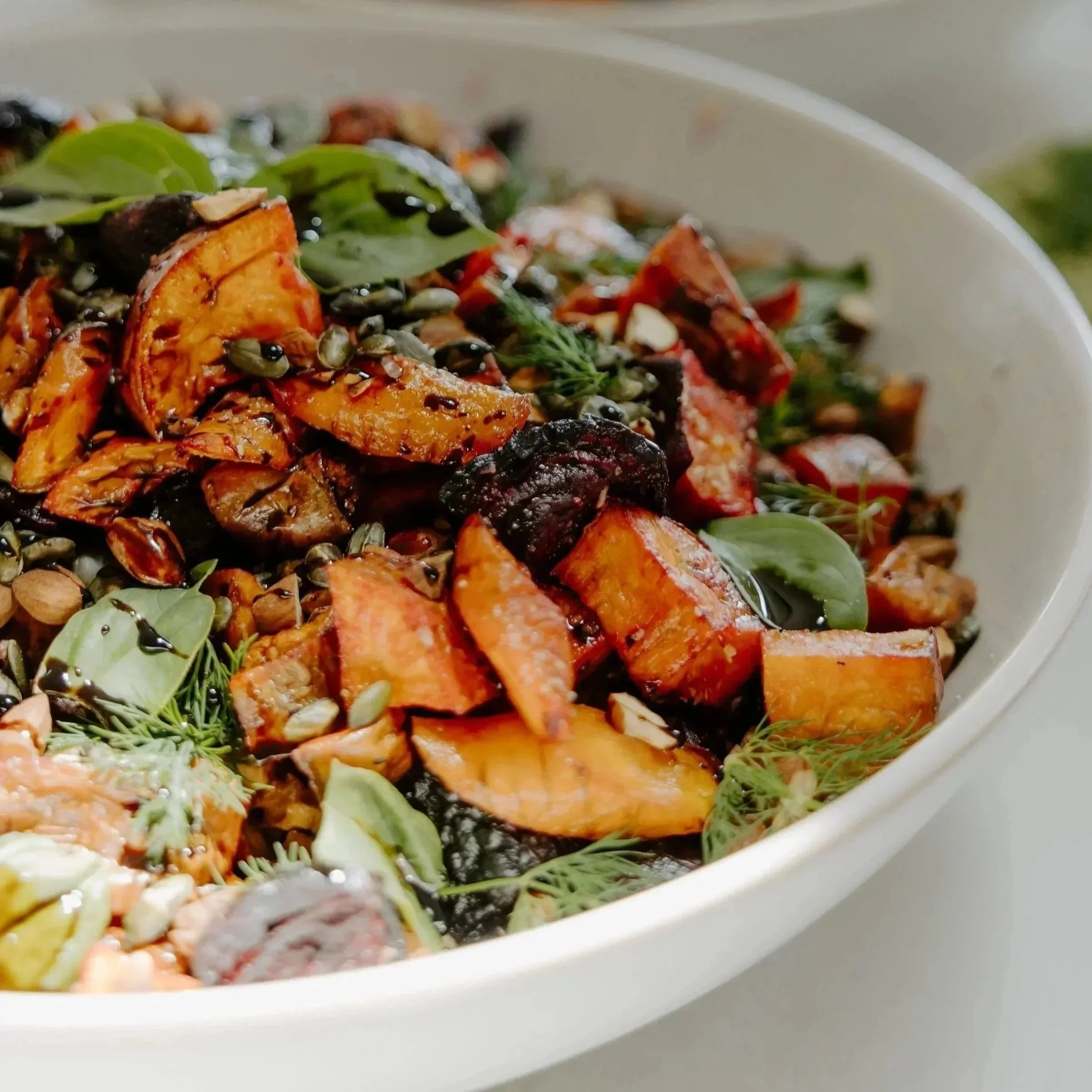 Close-up of a colorful salad in a white bowl, featuring roasted sweet potatoes, leafy greens, seeds, and a drizzle of dark dressing.