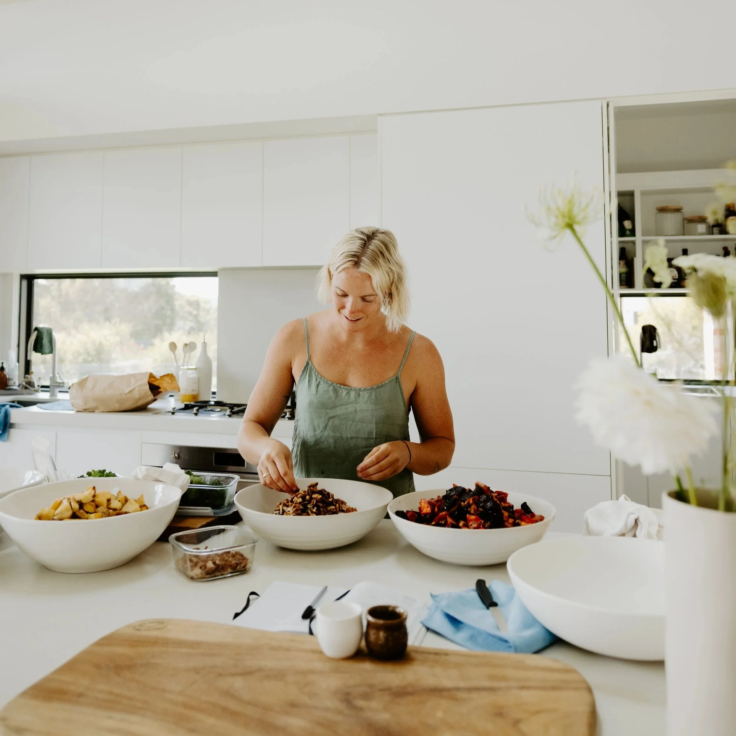 A woman with blonde hair wearing a green tank top is preparing food in a bright, modern kitchen. Various bowls of chopped vegetables and ingredients are spread out on the white countertop. The kitchen has large windows, white cabinets, and some decorations on the shelves.