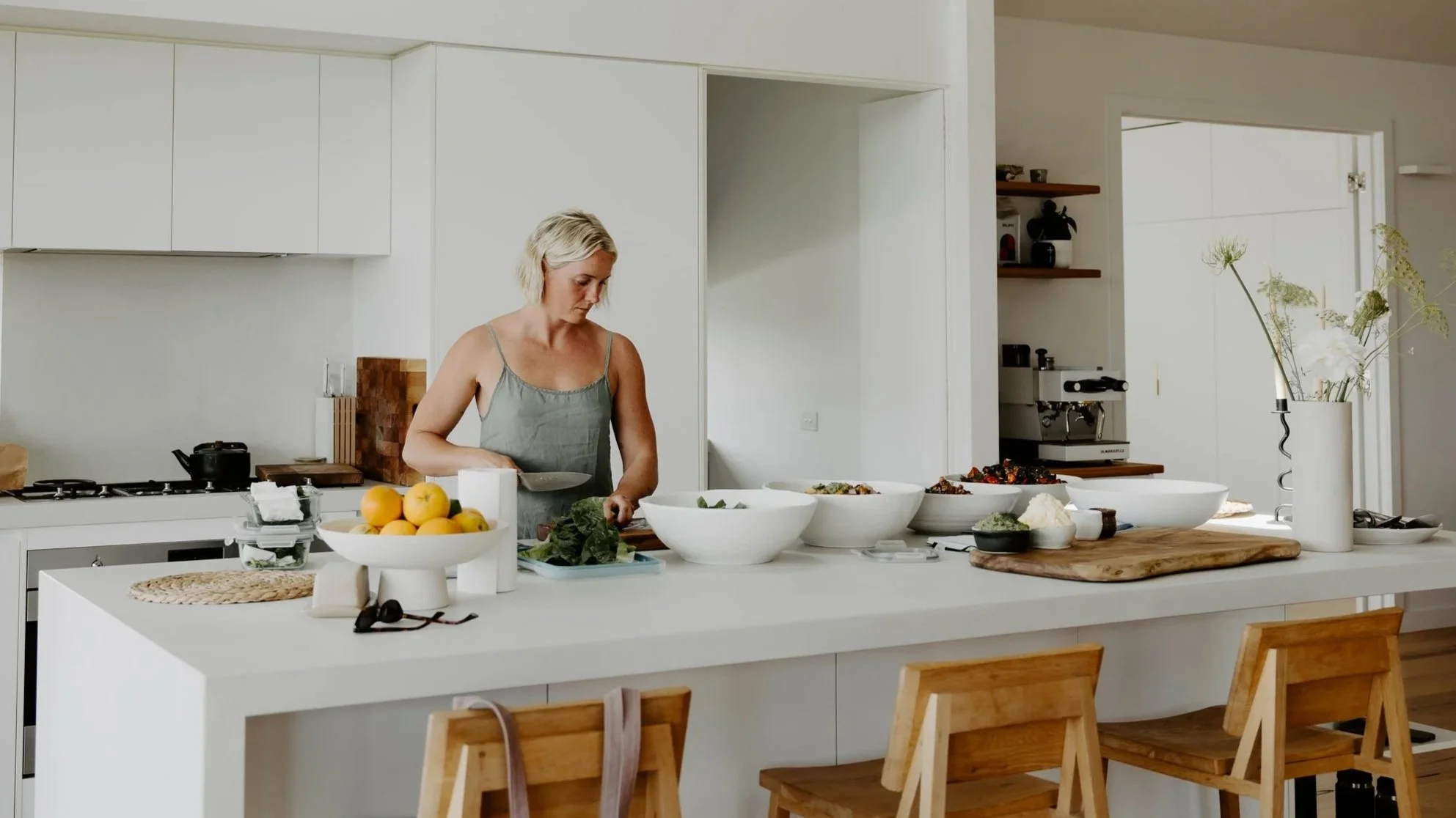 A woman with short blond hair wearing a gray sleeveless top prepares food at a kitchen island with several bowls of ingredients, a cutting board, and a vase with flowers in a modern, minimalist kitchen.