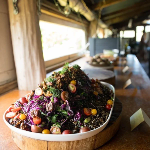 A colorful salad with various vegetables and toppings on a white dish placed on a wooden surface, inside a rustic setting with wooden walls and a window in the background.