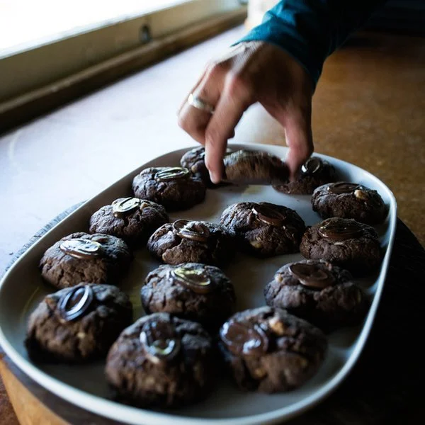 A hand reaching for a chocolate cookie from a white tray filled with chocolate cookies, on a wooden surface.