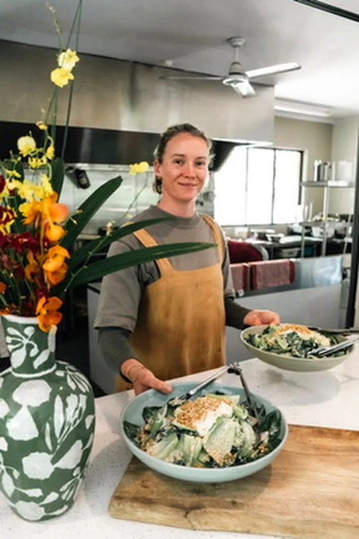 A woman standing in a kitchen holding two bowls of salad, with a large green and white patterned vase of flowers on the counter beside her.