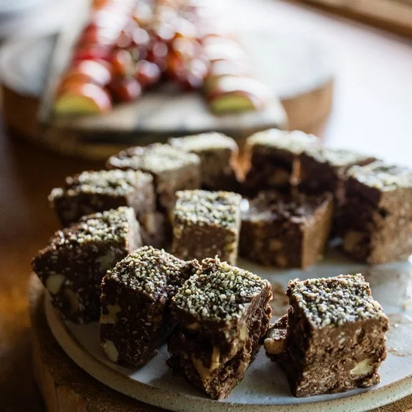 Close-up of chocolate brownie squares with nuts on a white plate, with blurred dessert in the background.
