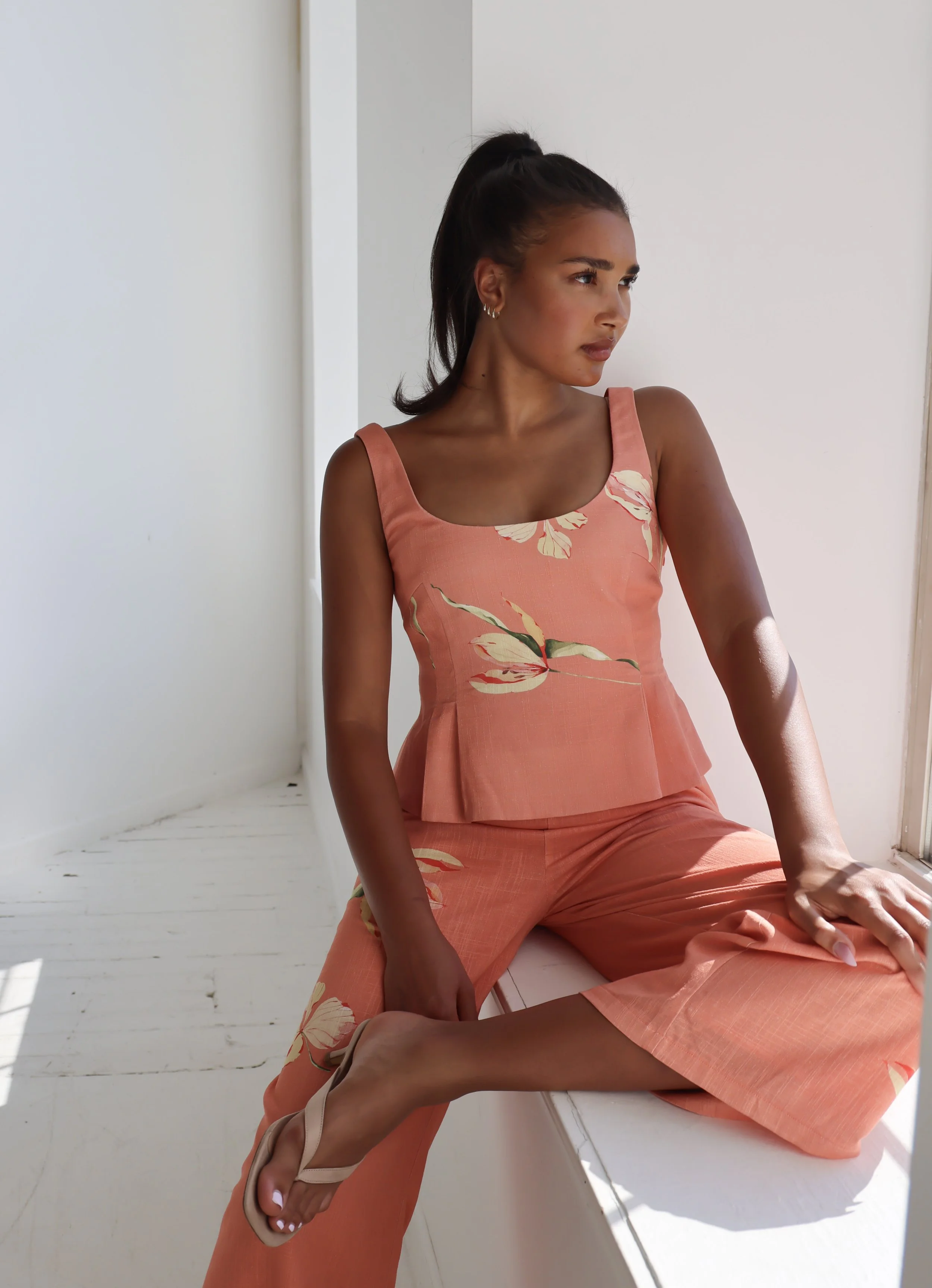 A woman seated on a white windowsill, wearing a coral-colored sleeveless dress with a floral pattern. She has dark hair tied back and is wearing earrings. She is looking to her left with sunlight illuminating her face and arm, and wearing beige heels.