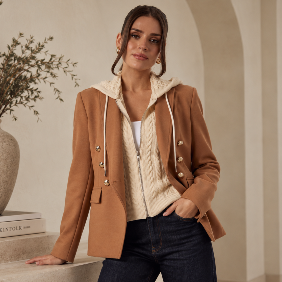 A woman wearing a tan blazer over a cream cable-knit sweater with a hood, standing indoors near a table with a large vase and a book. She has dark hair and is posing with her left hand in her pocket.