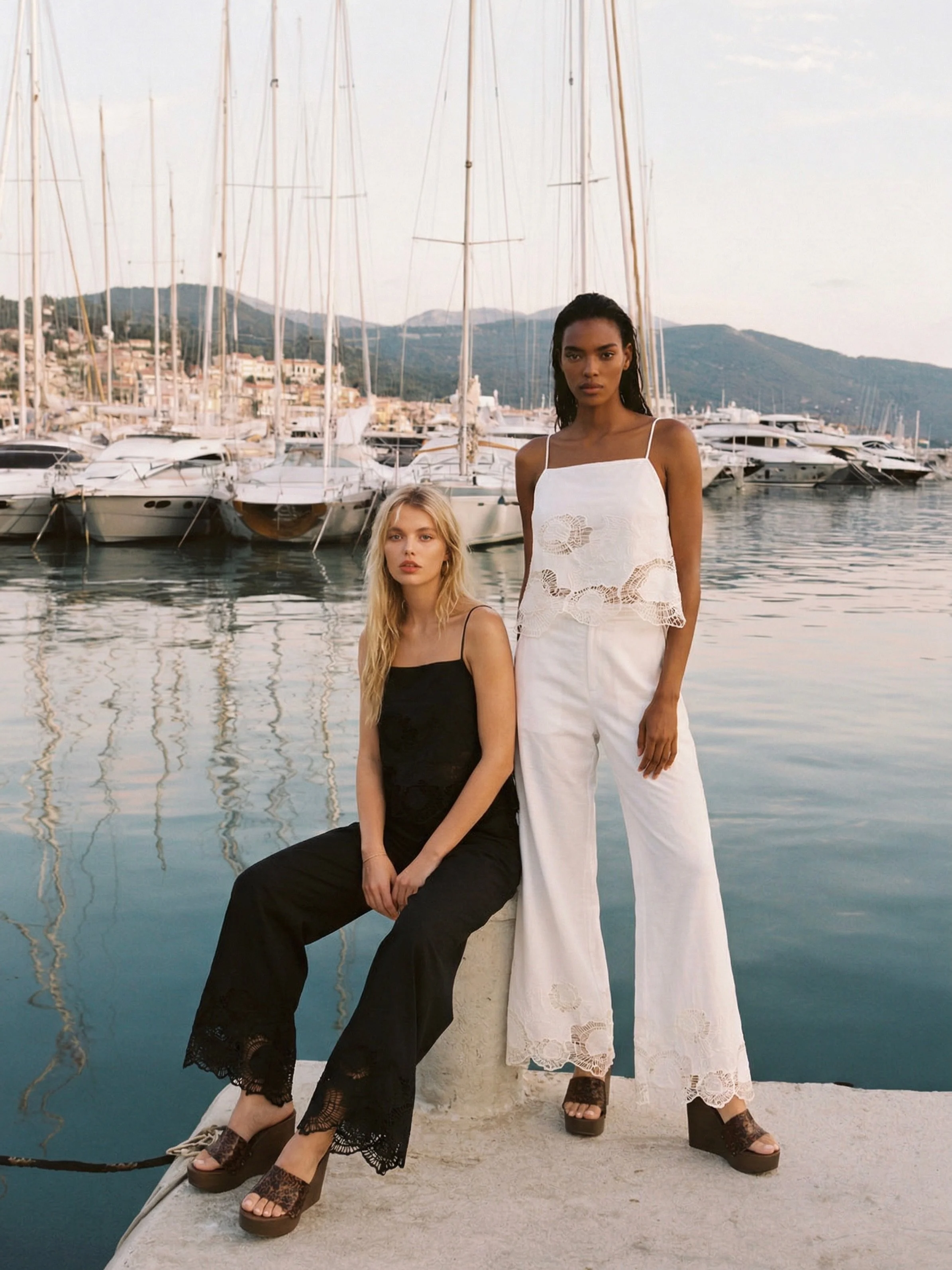Two women near a marina with several sailboats and yachts in the background, standing on a dock or pier with mountains in the distance.