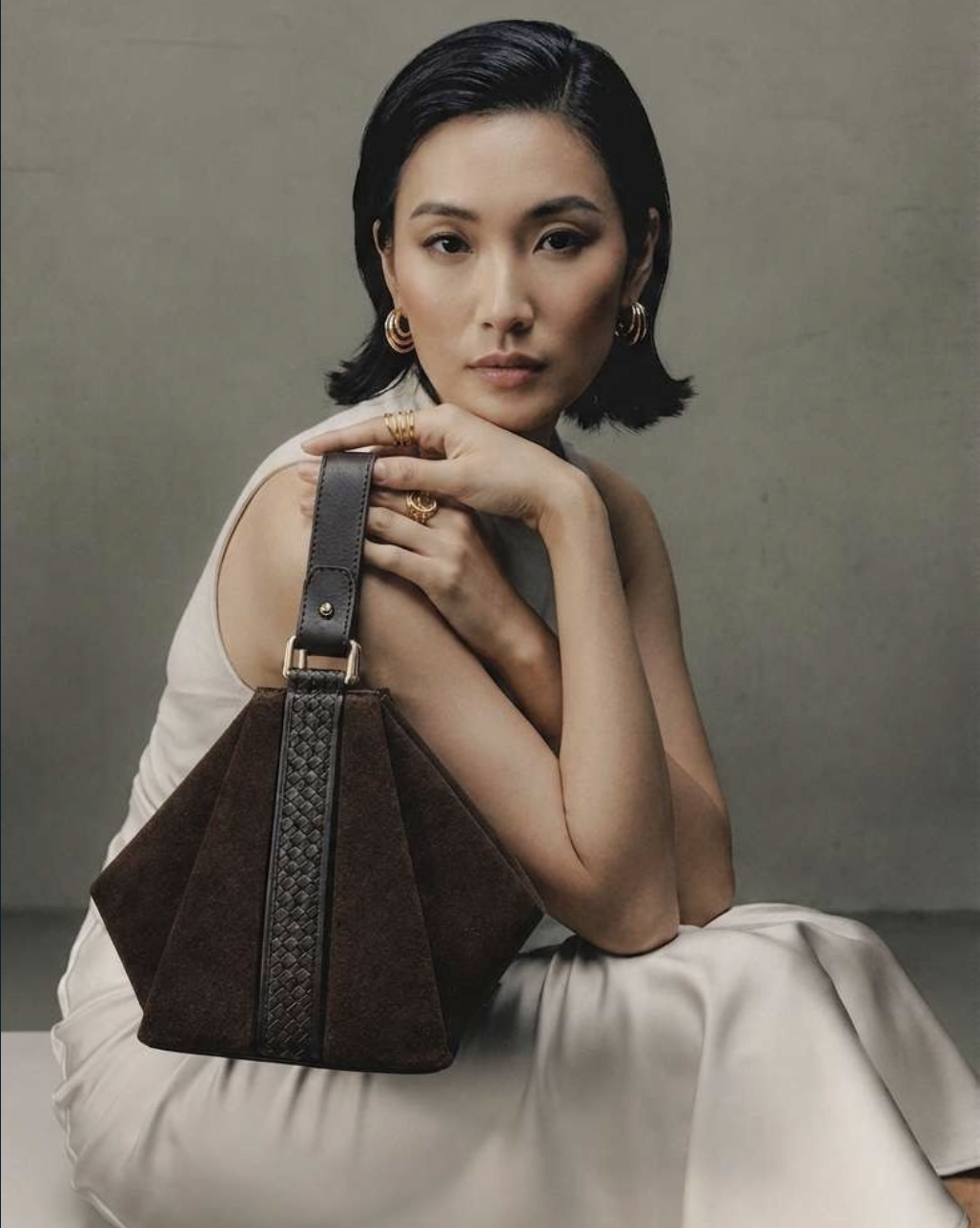 A woman with short black hair wearing gold hoop earrings, gold rings, and a sleeveless light-colored dress, sitting against a plain gray background with a dark brown handbag resting on her lap.
