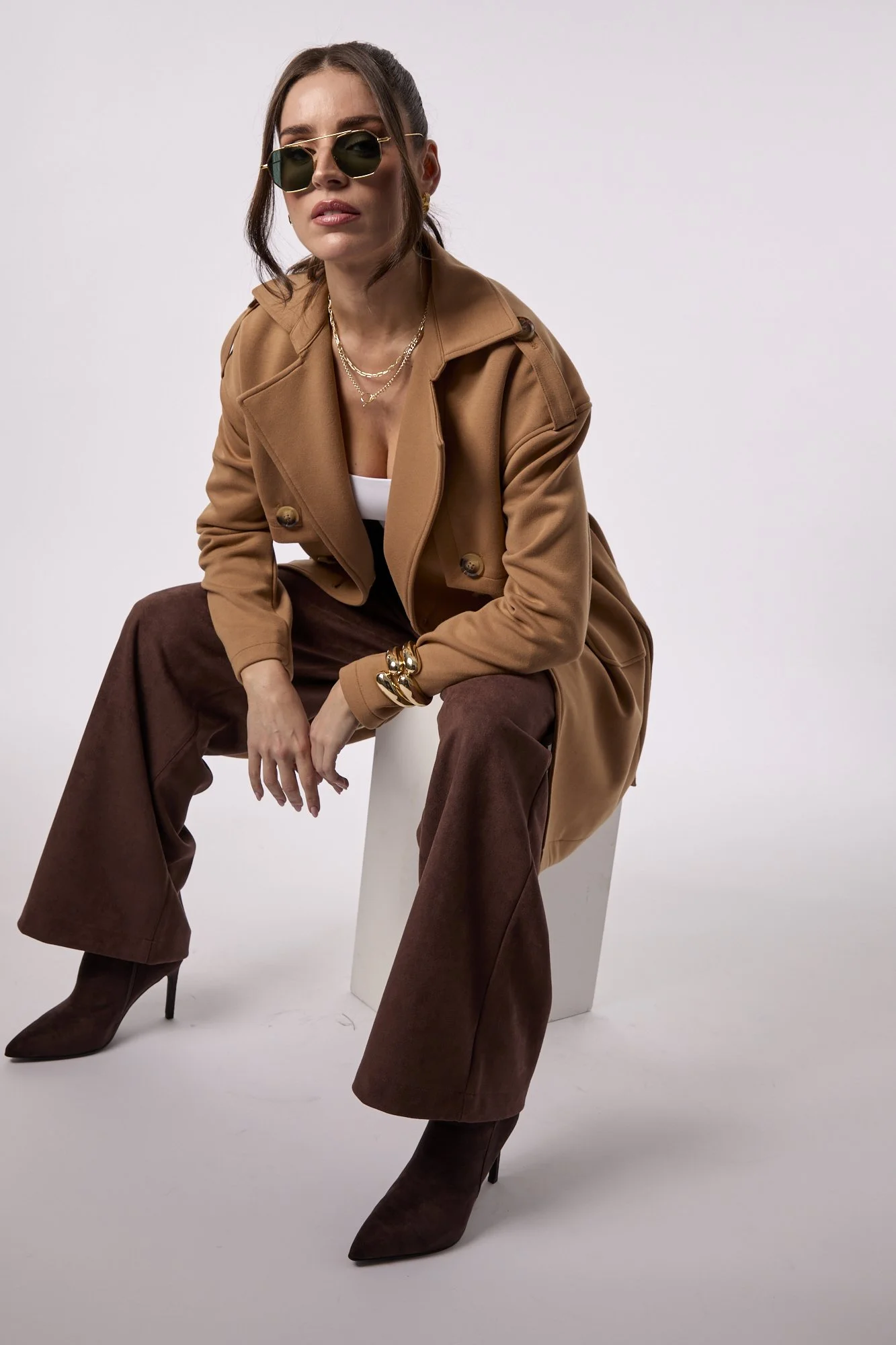 A woman in a tan trench coat, white top, brown wide-leg pants, and brown high-heeled boots, sitting on a white stool against a plain background. She is wearing dark glasses, layered gold necklaces, and gold bracelets, with her hair styled in loose waves.