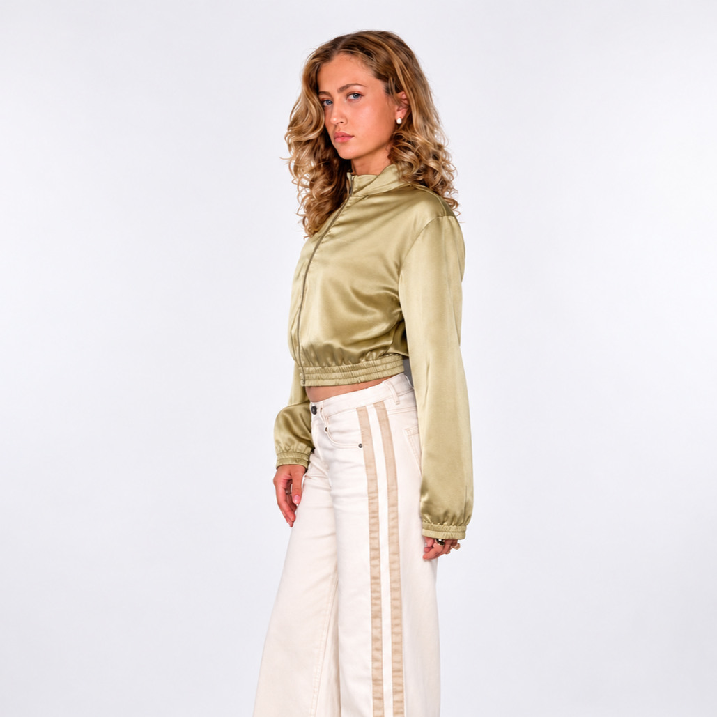 A woman in a tan trench coat, white top, brown wide-leg pants, and brown high-heeled boots, sitting on a white stool against a plain background. She is wearing dark glasses, layered gold necklaces, and gold bracelets, with her hair styled in loose waves.