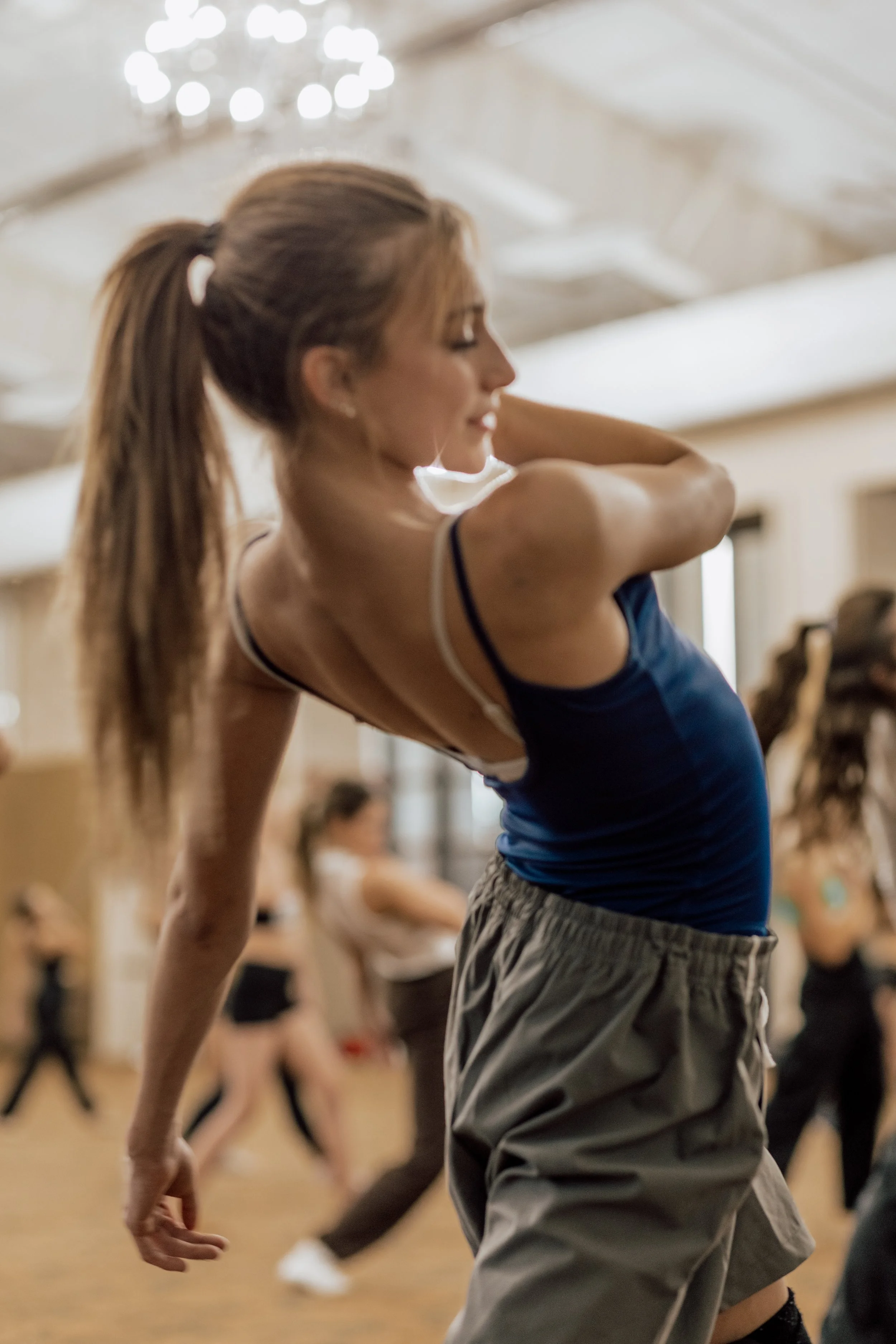 A woman with a ponytail stretching or warming up in a dance or fitness class, with other participants blurred in the background.