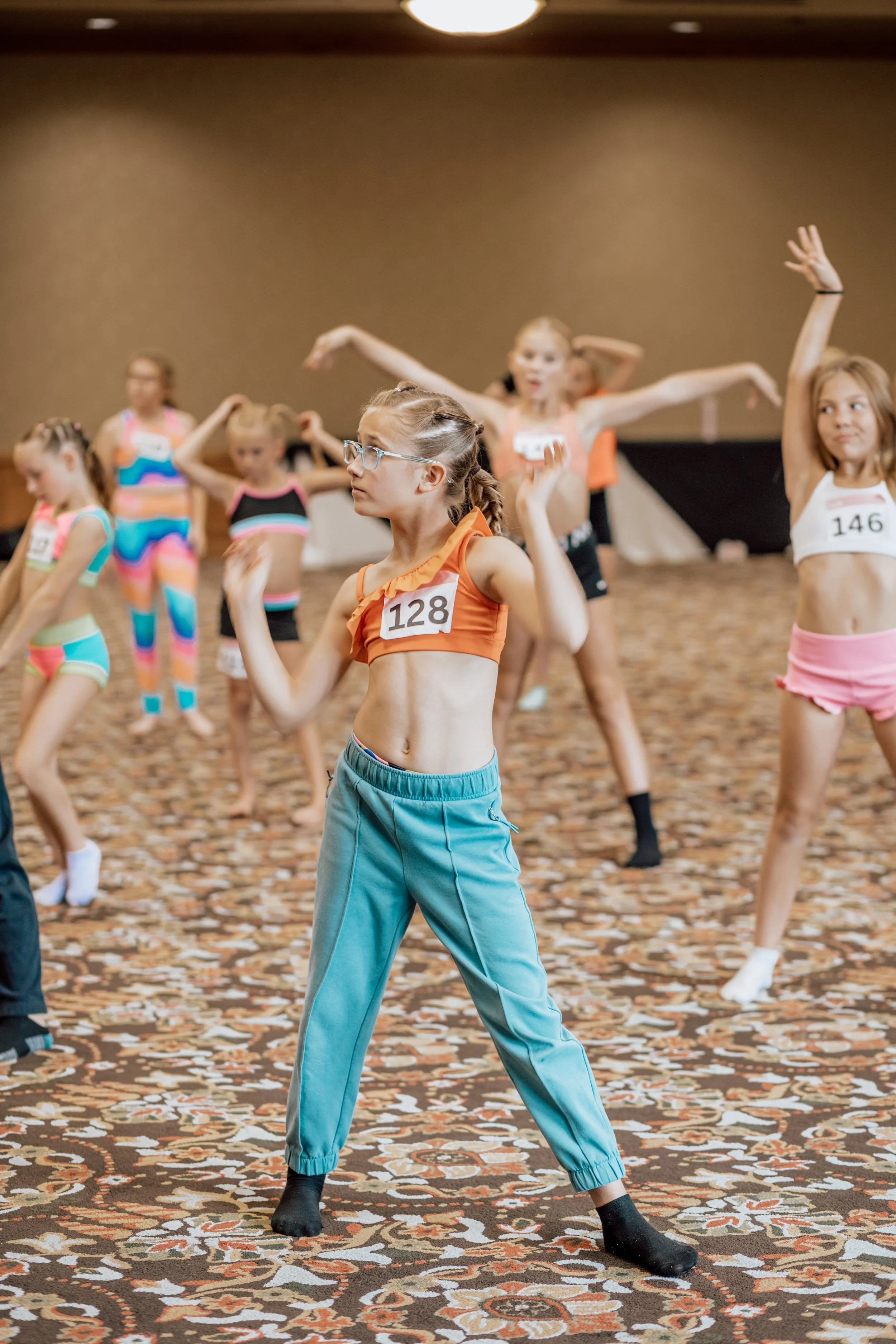 Young girls practicing dance moves in colorful athletic wear during a dance class or rehearsal in a large indoor space.