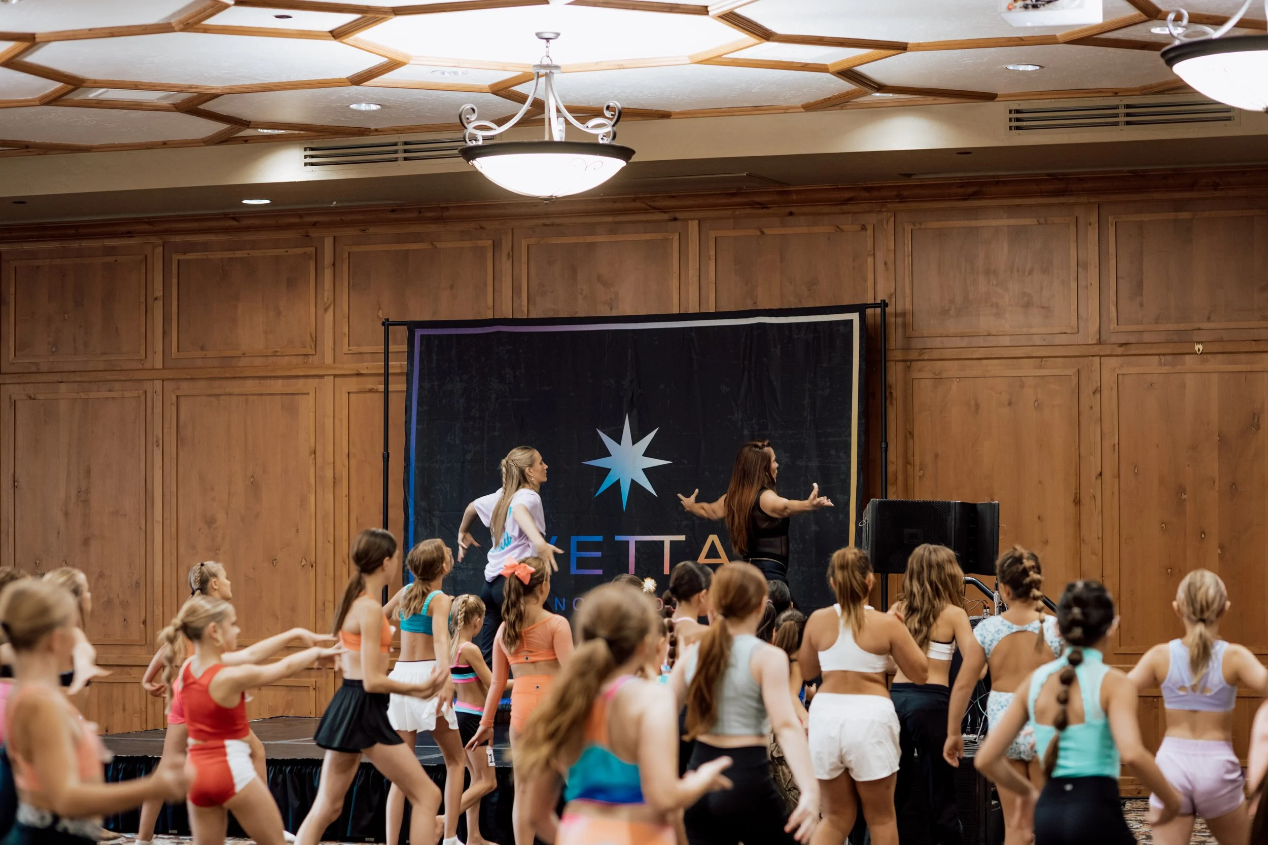 Young girls participating in a dance or gymnastics class in a large indoor space with wood-paneled walls and an elevated stage at the front. There are banners and a black backdrop behind the stage, and ceiling lights overhead.