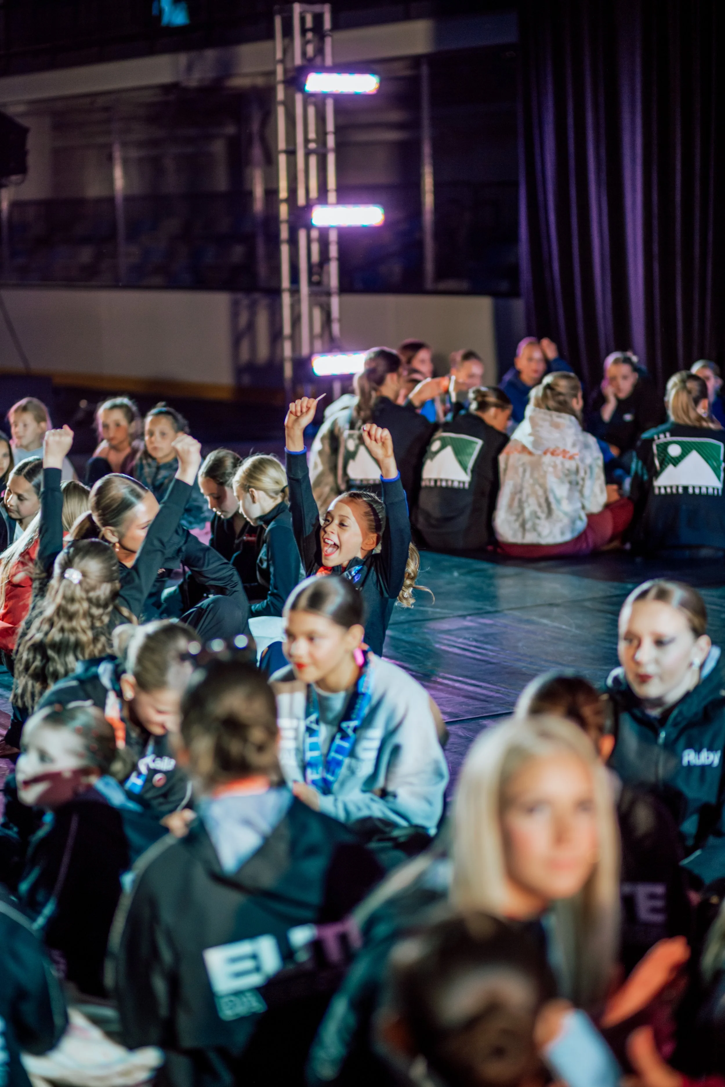 A group of young girls sitting on the floor of a stage, some raising their hands and cheering, in a dimly lit room with purple lights and dark curtains in the background. Vetta Dance Project Expo