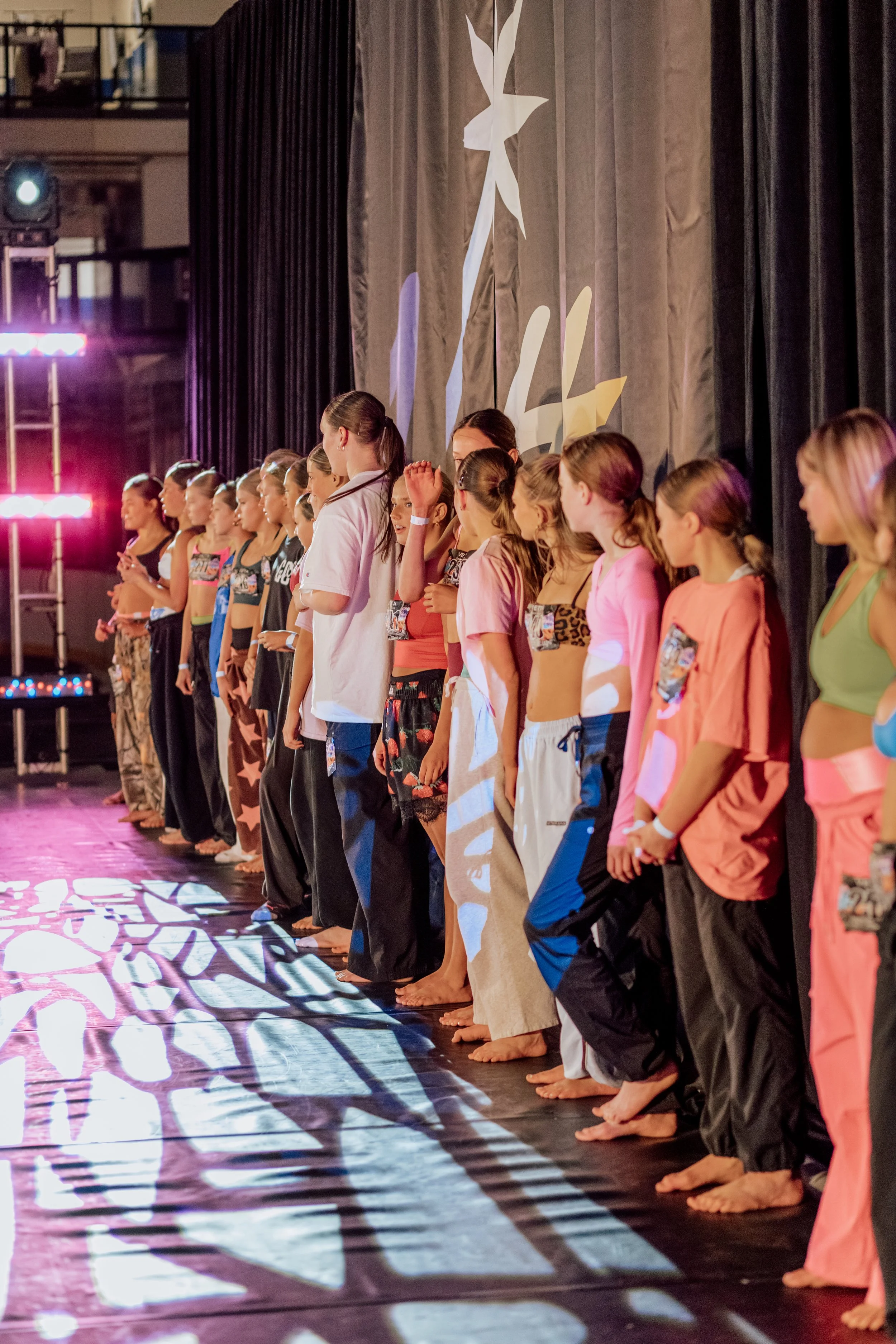 Line of young women in colorful workout attire standing barefoot on a stage, preparing for a performance, with a curtain and stage lights in the background.