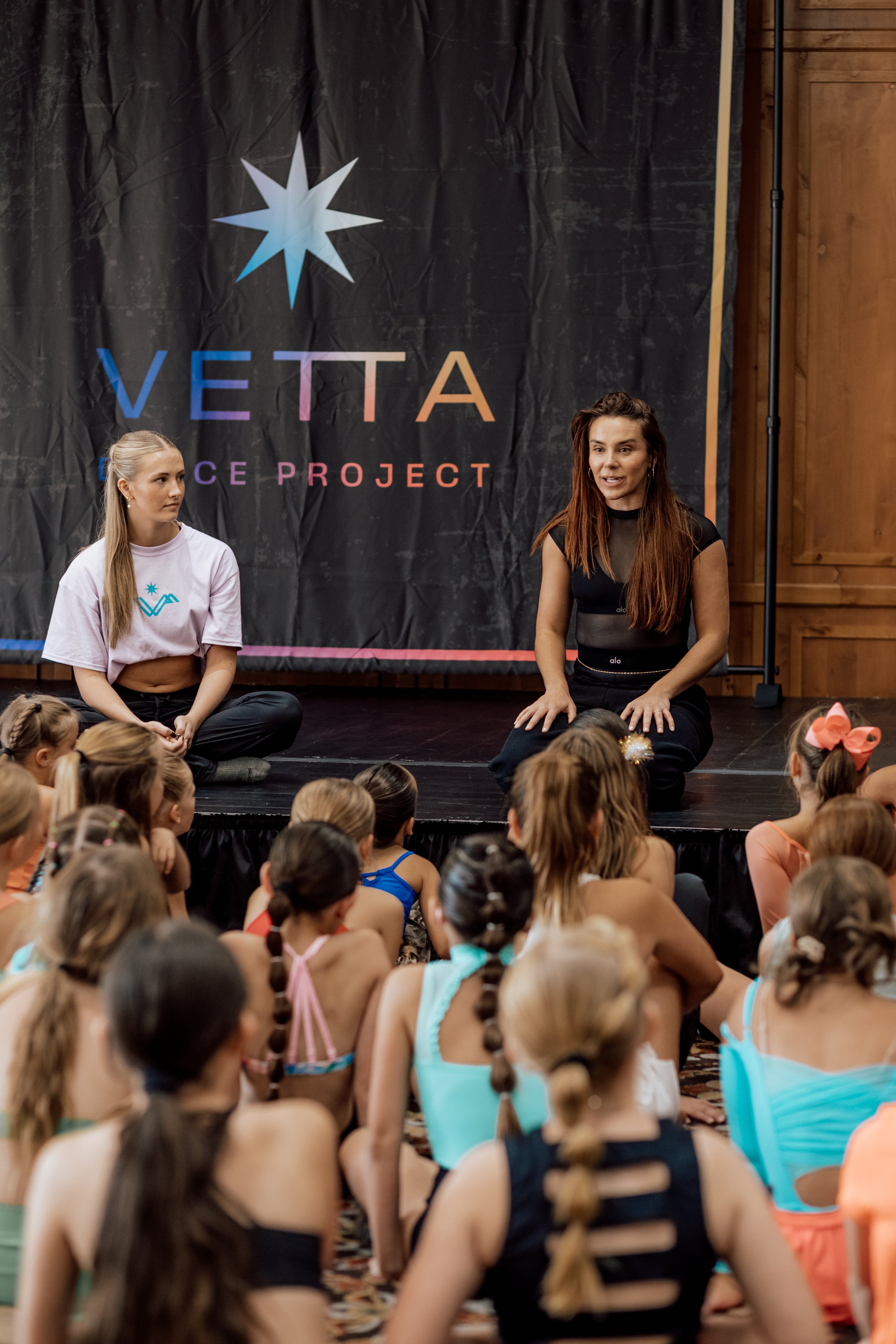 Young woman with long brown hair, wearing black dancewear, sitting on stage in front of a black curtain with the 'VETTA Dance Project' logo, addressing a group of children in a dance studio.