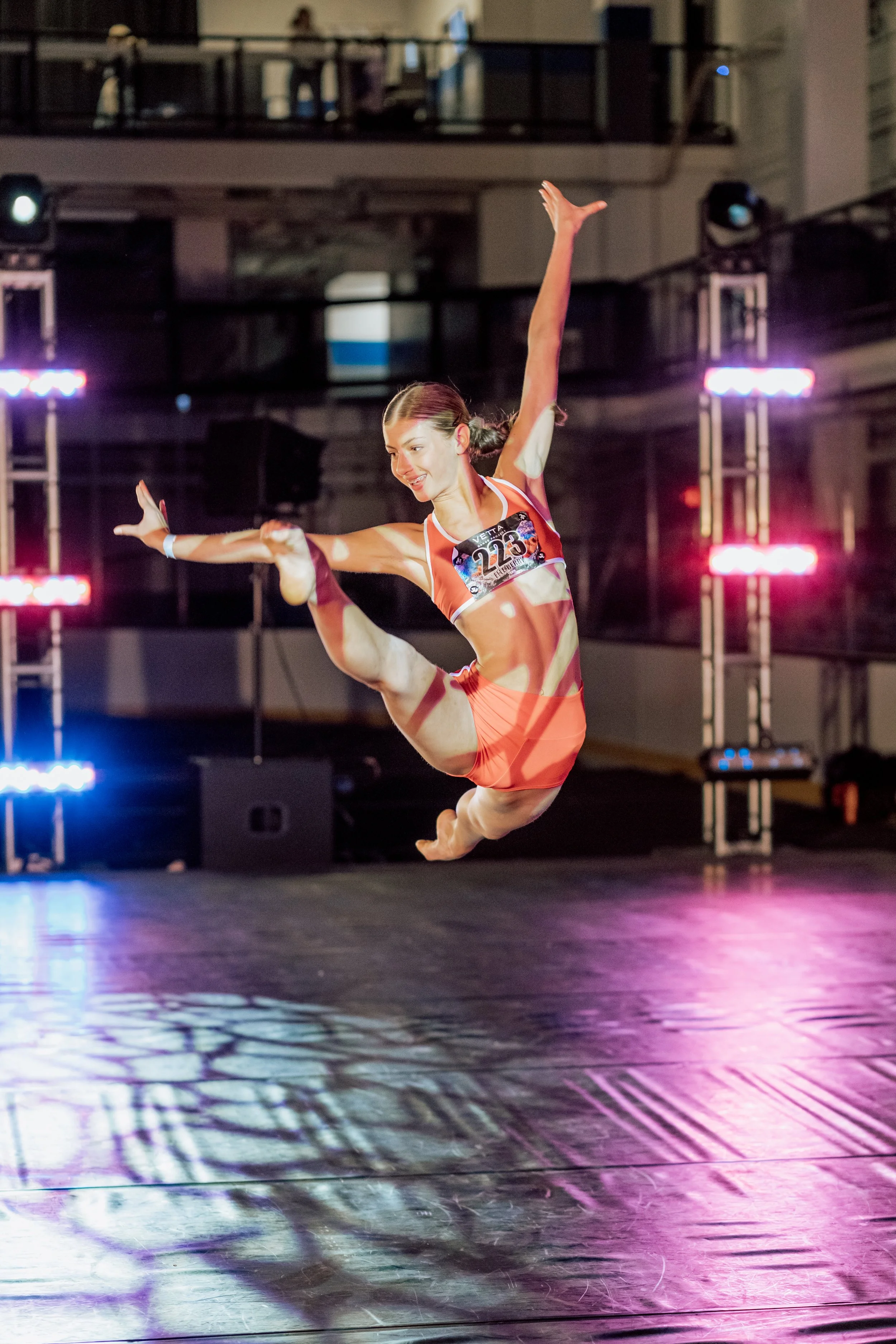 A female gymnast mid-performance, balancing on one leg with one arm extended and the other raised, in a brightly lit indoor sports arena.