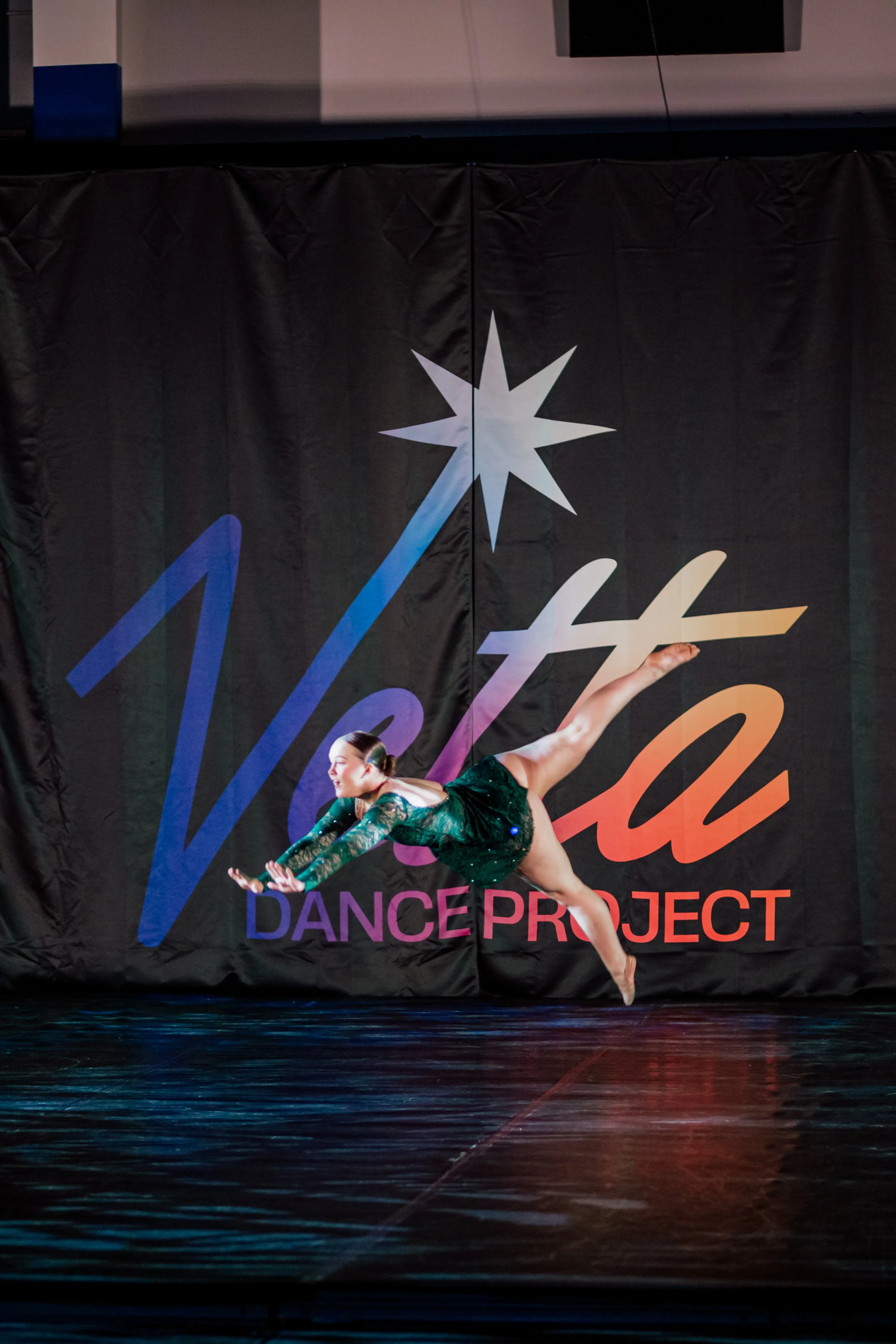 A dancer in a green costume performing a leap on stage in front of a backdrop with the words 'Dance Project' and a starburst logo.