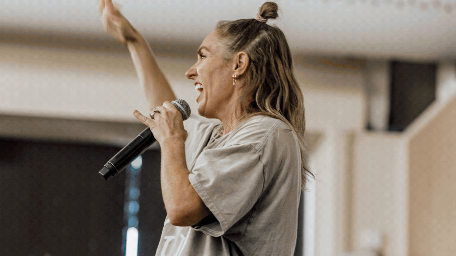 A woman with long, wavy hair in a partial ponytail, wearing earrings and a casual beige shirt, speaking into a microphone and gesturing with her right hand in an indoor setting. Vetta Dance Project Expo