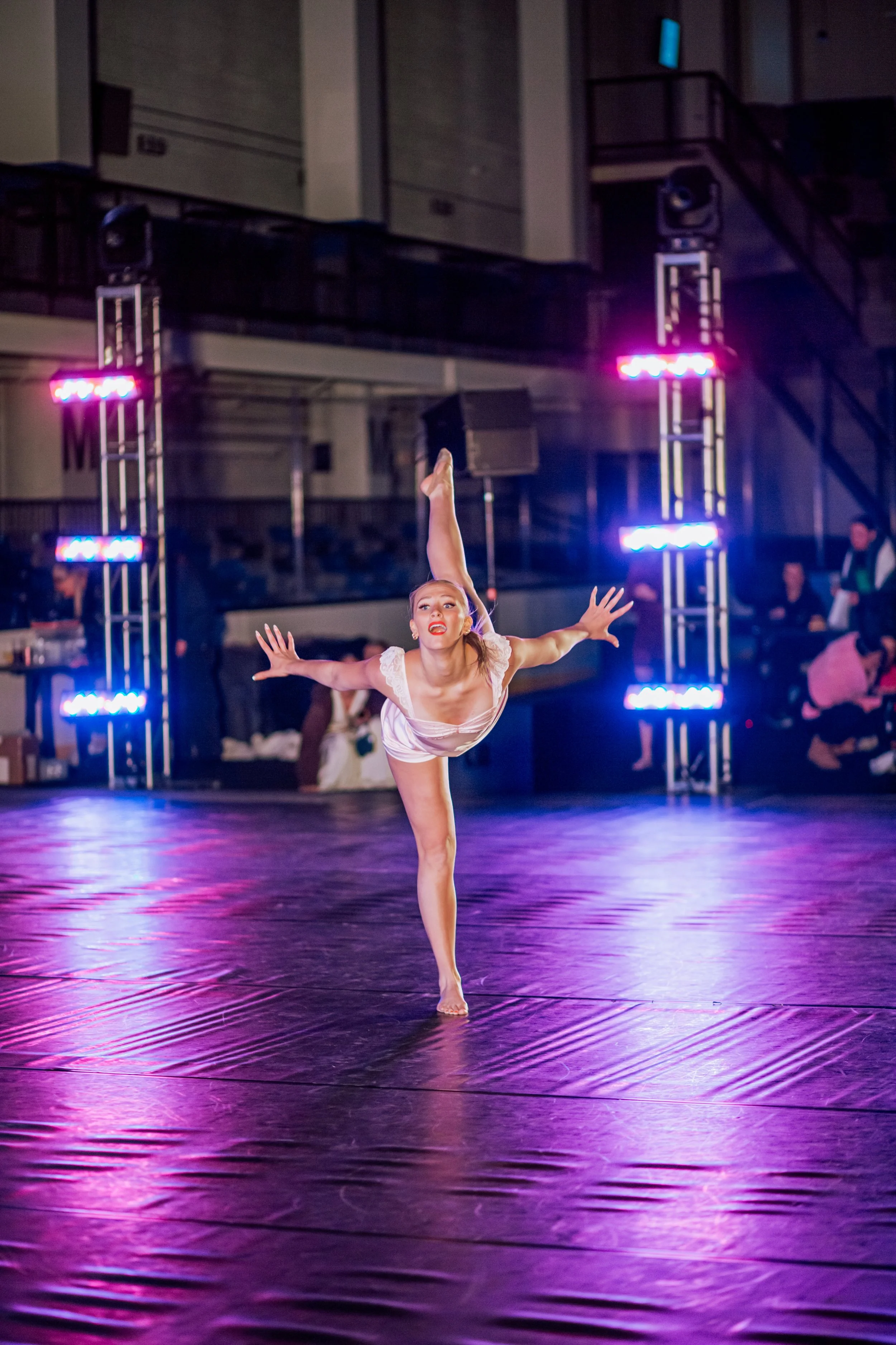 A female gymnast in a white costume performing an expressive dance move on a wooden floor during a nighttime indoor event with blue and purple stage lights.