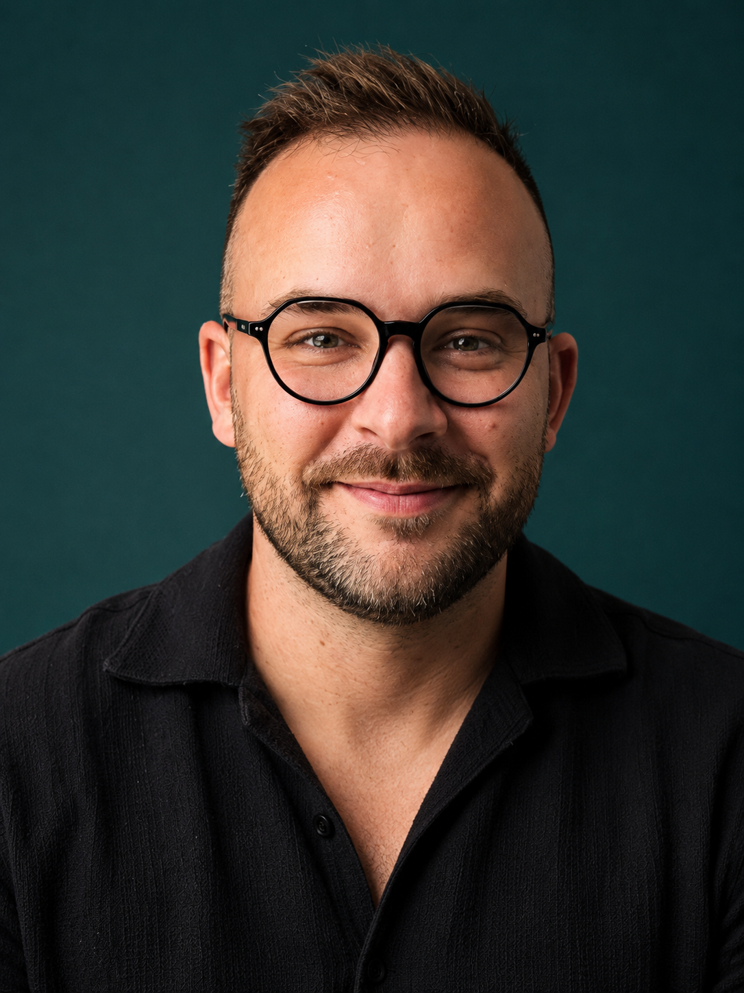 Portrait d'un homme souriant avec des lunettes rondes, barbe et cheveux courts, portant une chemise noire.
