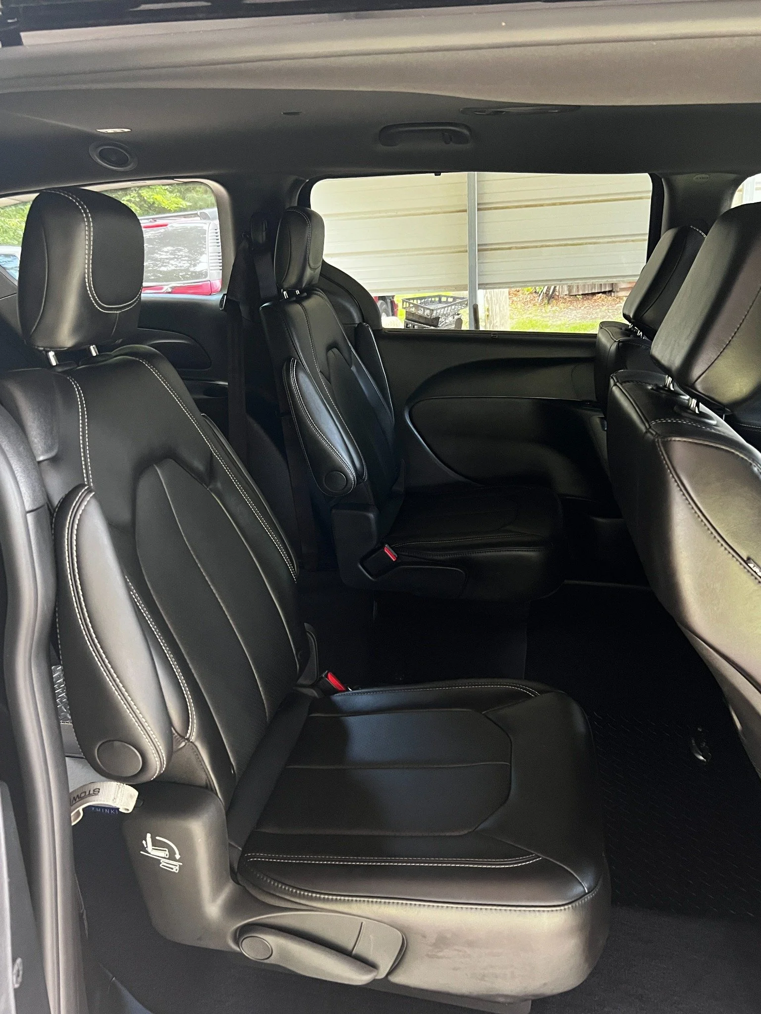 Interior of a black SUV showing leather seats, headrests, and the rear window with a view of the outside, including a partially open garage door, a plastic crate, and some greenery.